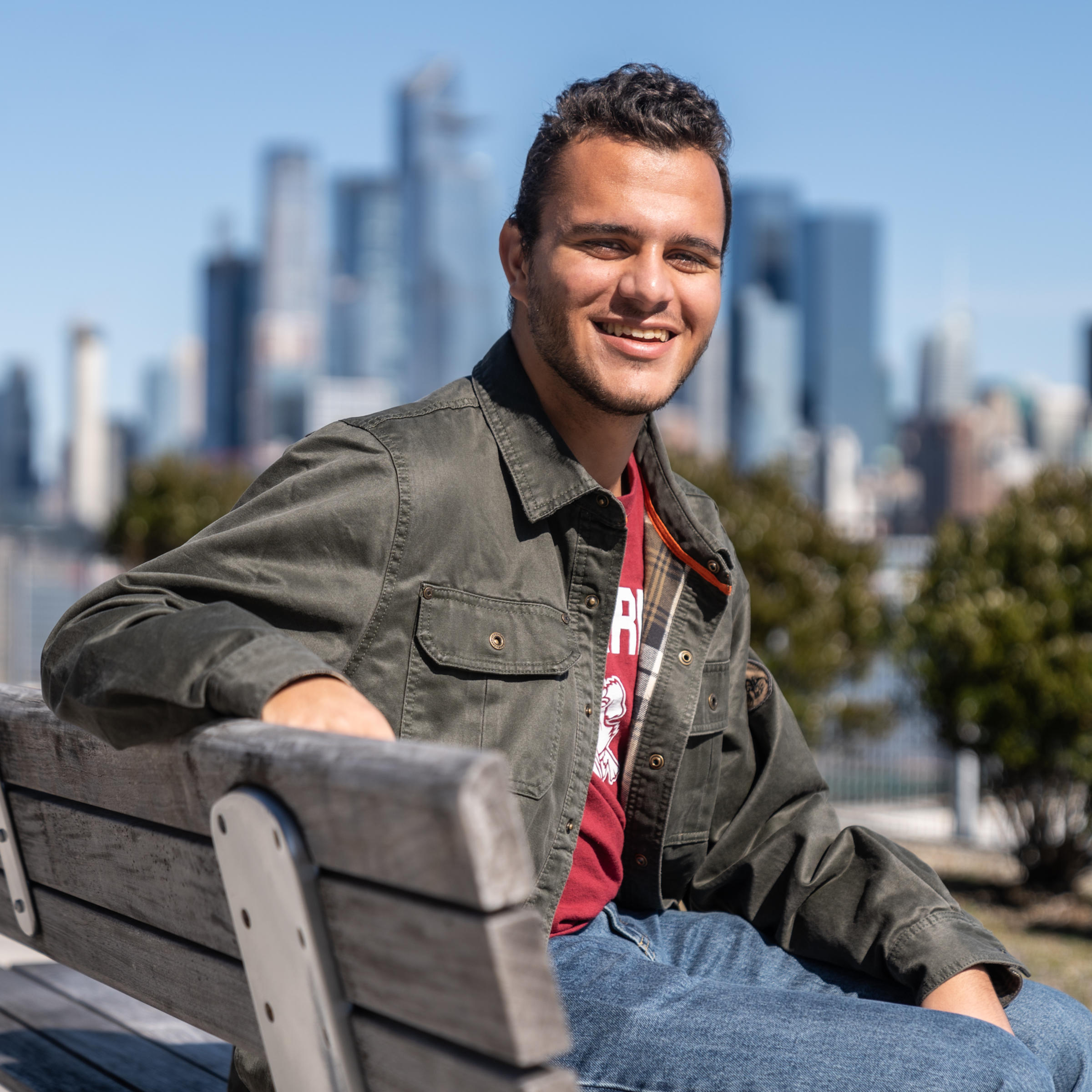 Gareth Maritz '23 sits on a bench with part of the New York City skyline in view behind him on a sunny day.