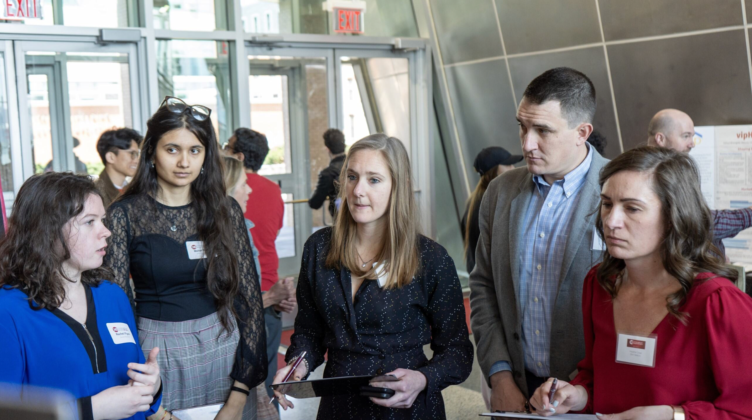 A competition judge, wearing a black dress, stands in the middle of a group of four students as they explain their project.