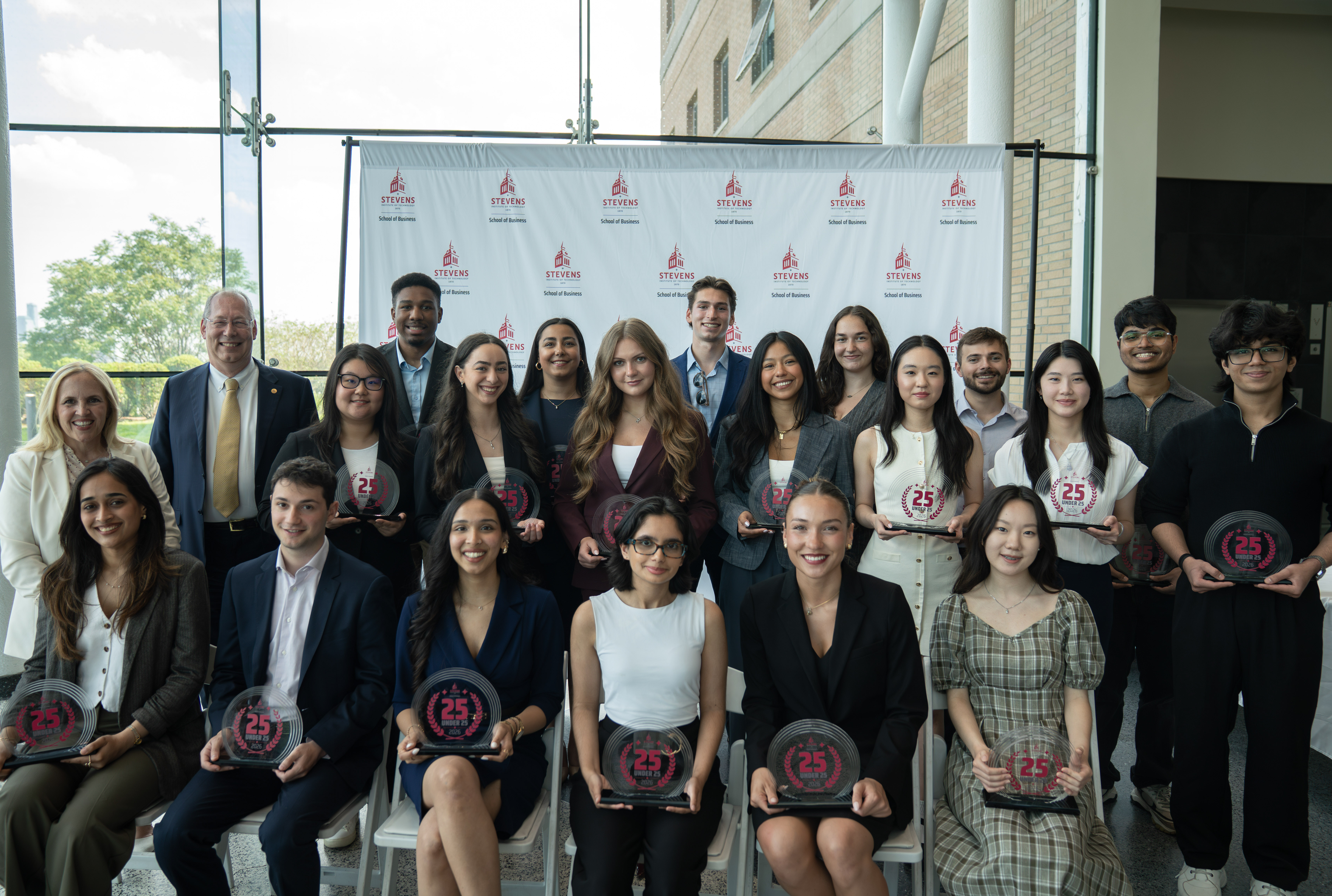 Inaugural 25 Under 25 cohort members pose for a group photo. They are dressed in business attire and are posed in front of a School of Business step and repeat banner