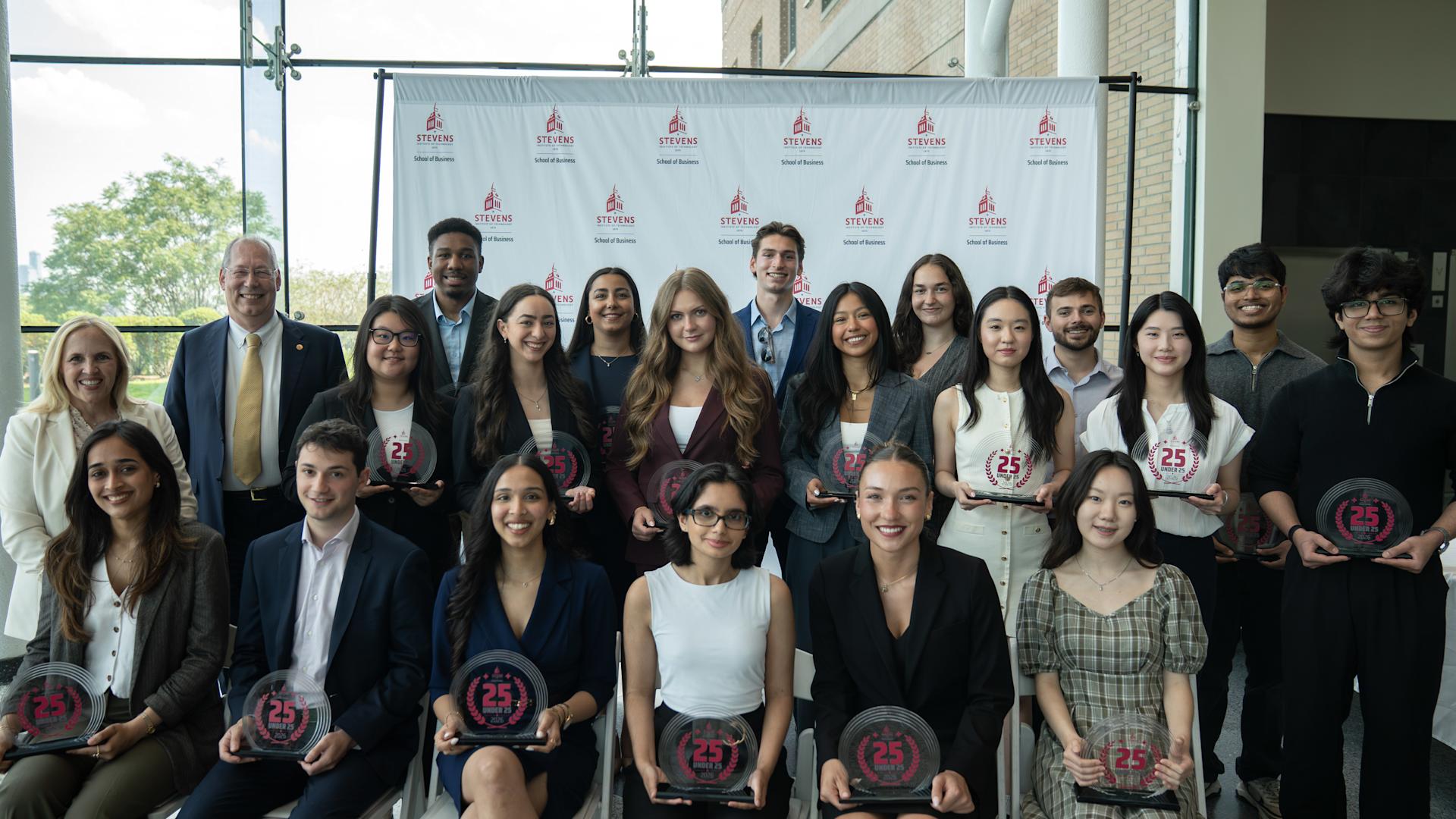 Inaugural 25 Under 25 cohort members pose for a group photo. They are dressed in business attire and are posed in front of a School of Business step and repeat banner