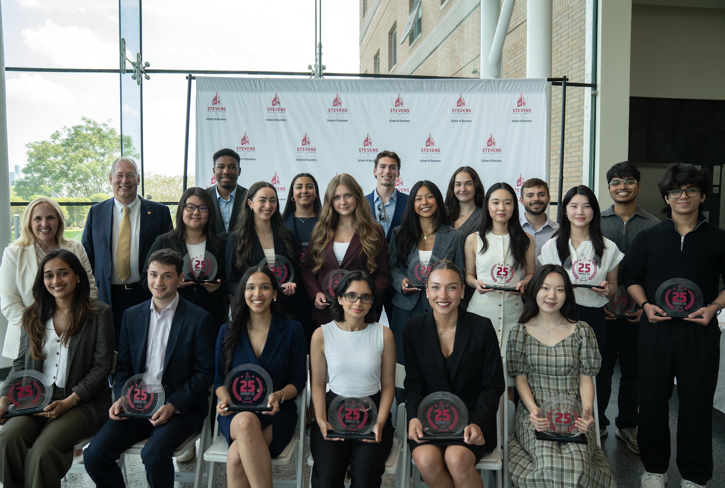 Inaugural 25 Under 25 cohort members pose for a group photo. They are dressed in business attire and are posed in front of a School of Business step and repeat banner