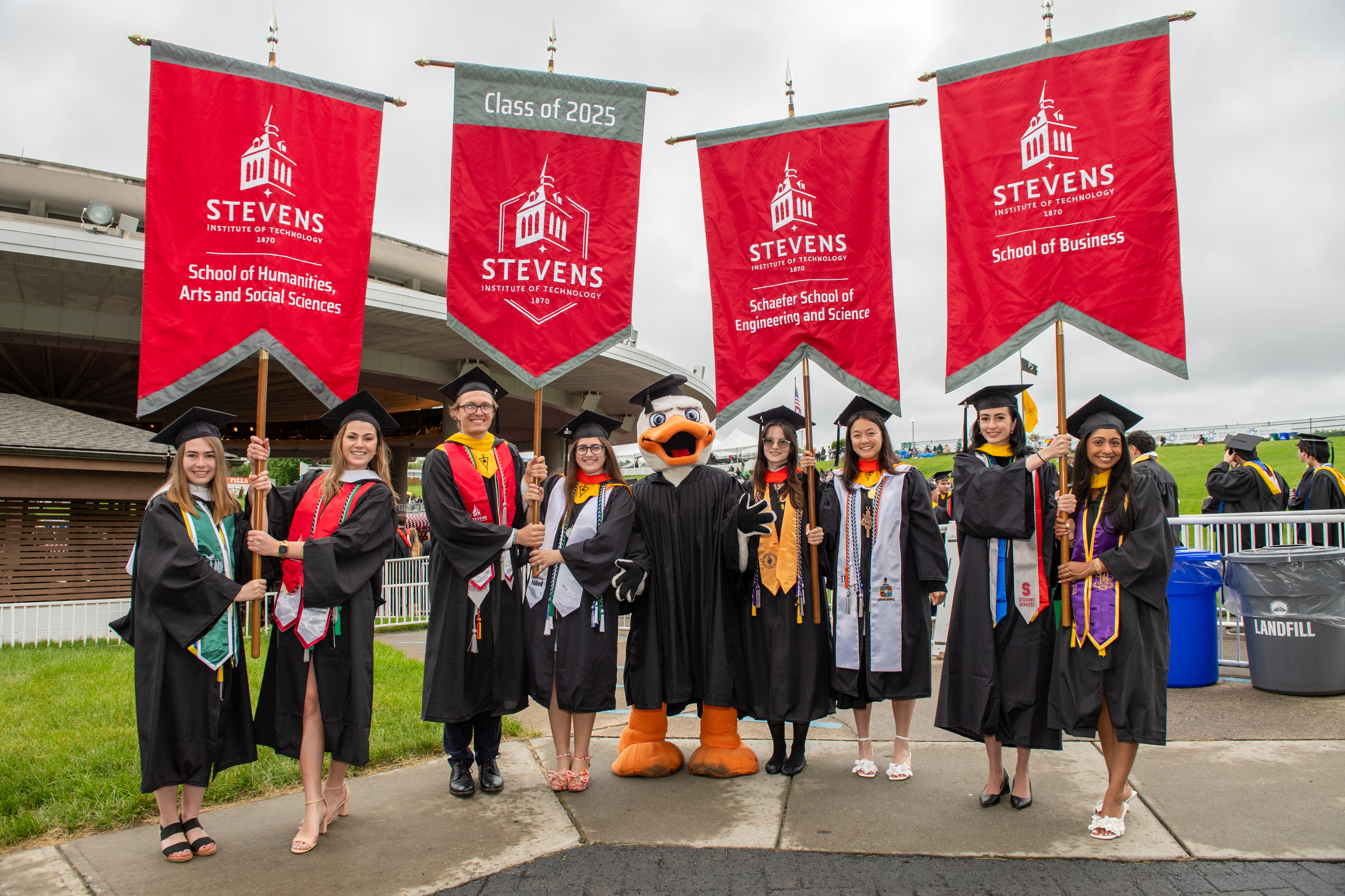 Students dressed in commencement regalia from each school hold the Class of 2025 banners.