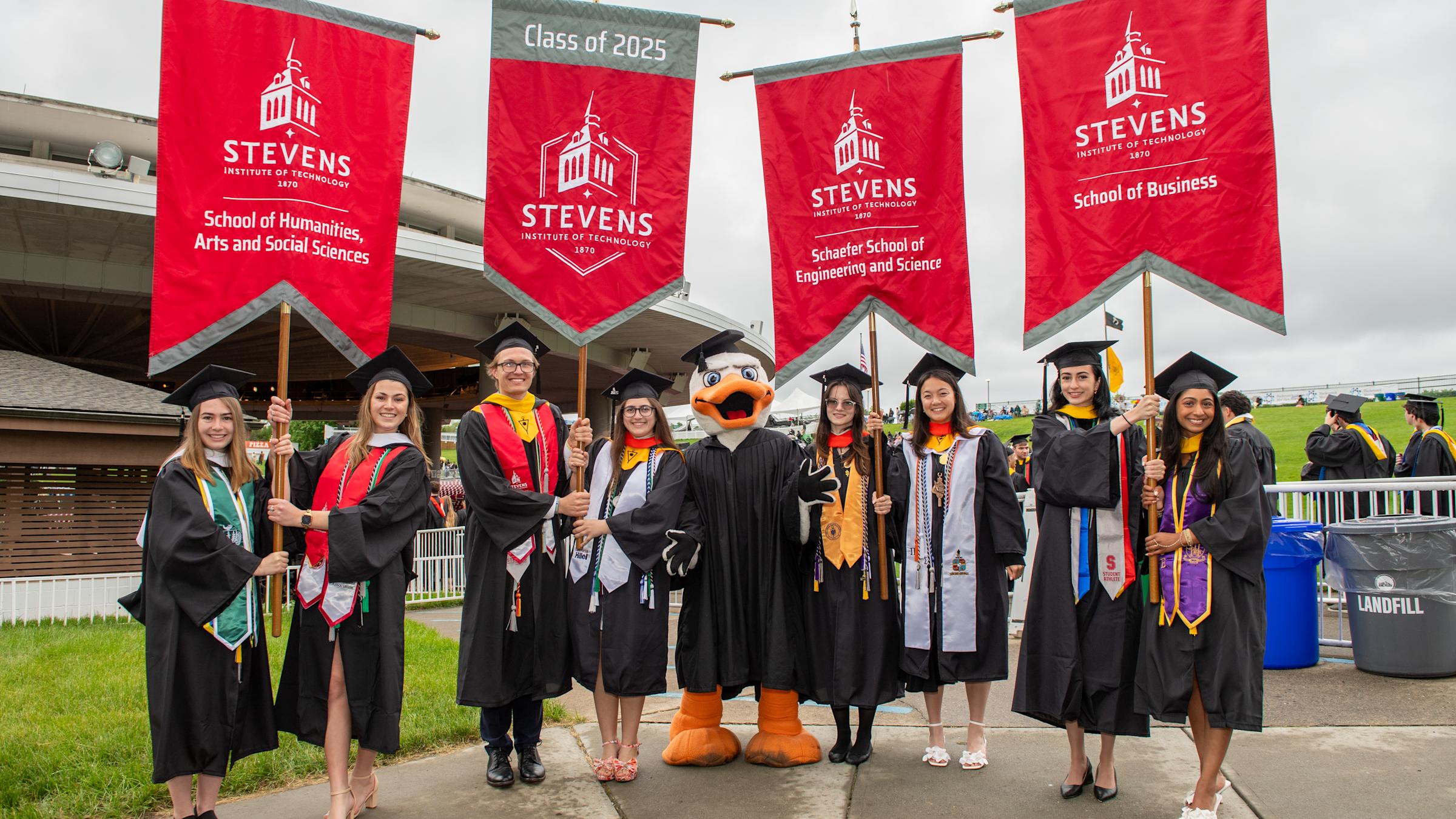 Students dressed in commencement regalia from each school hold the Class of 2025 banners.