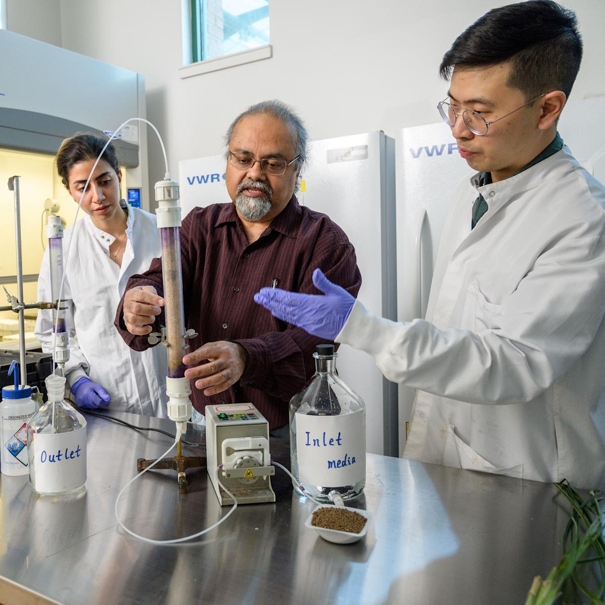 Stevens professor Dibs Sarkar is flanked by two students at a lab workstation with a test tube and glass bottles.