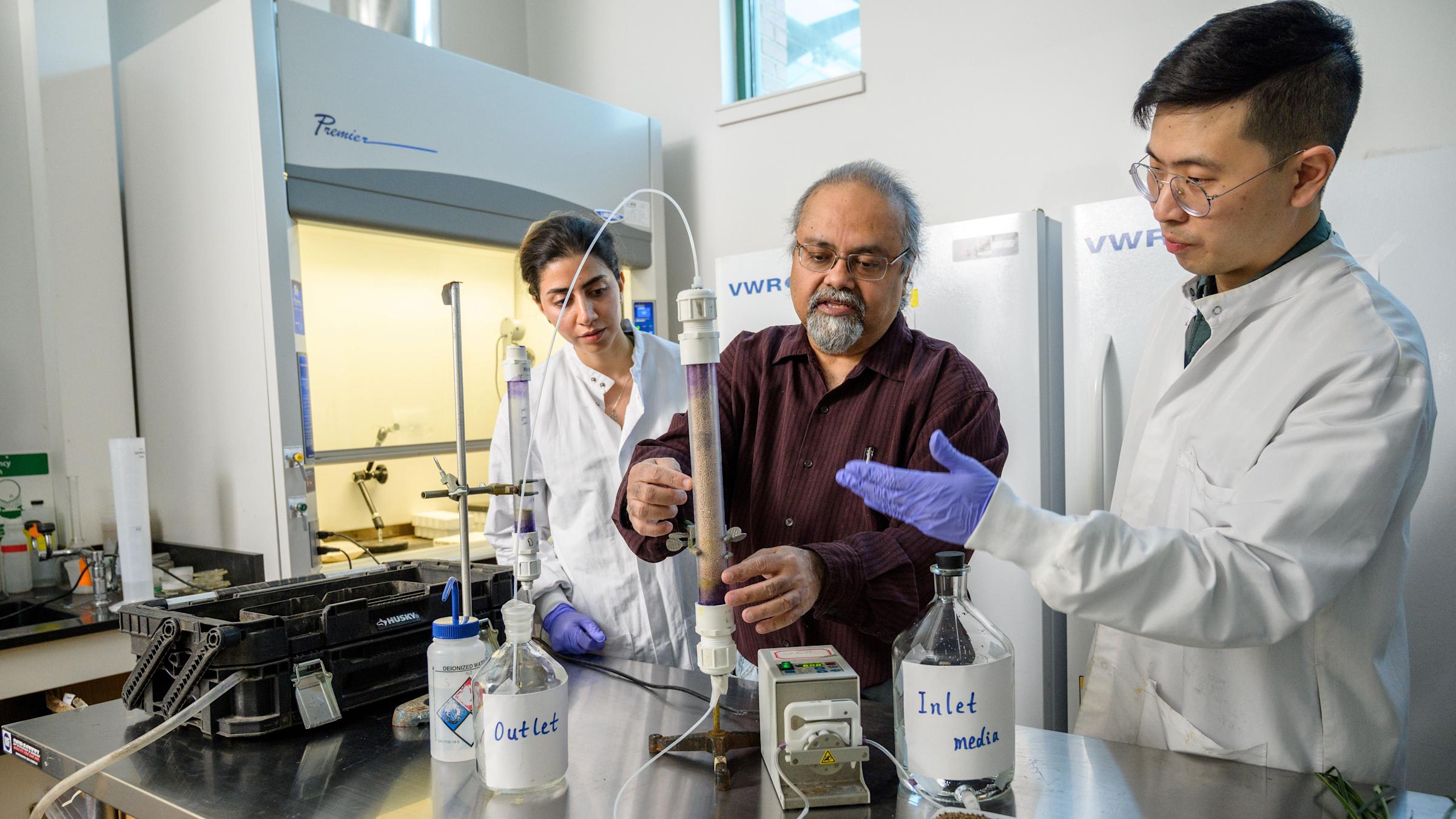 Stevens professor Dibs Sarkar is flanked by two students at a lab workstation with a test tube and glass bottles.