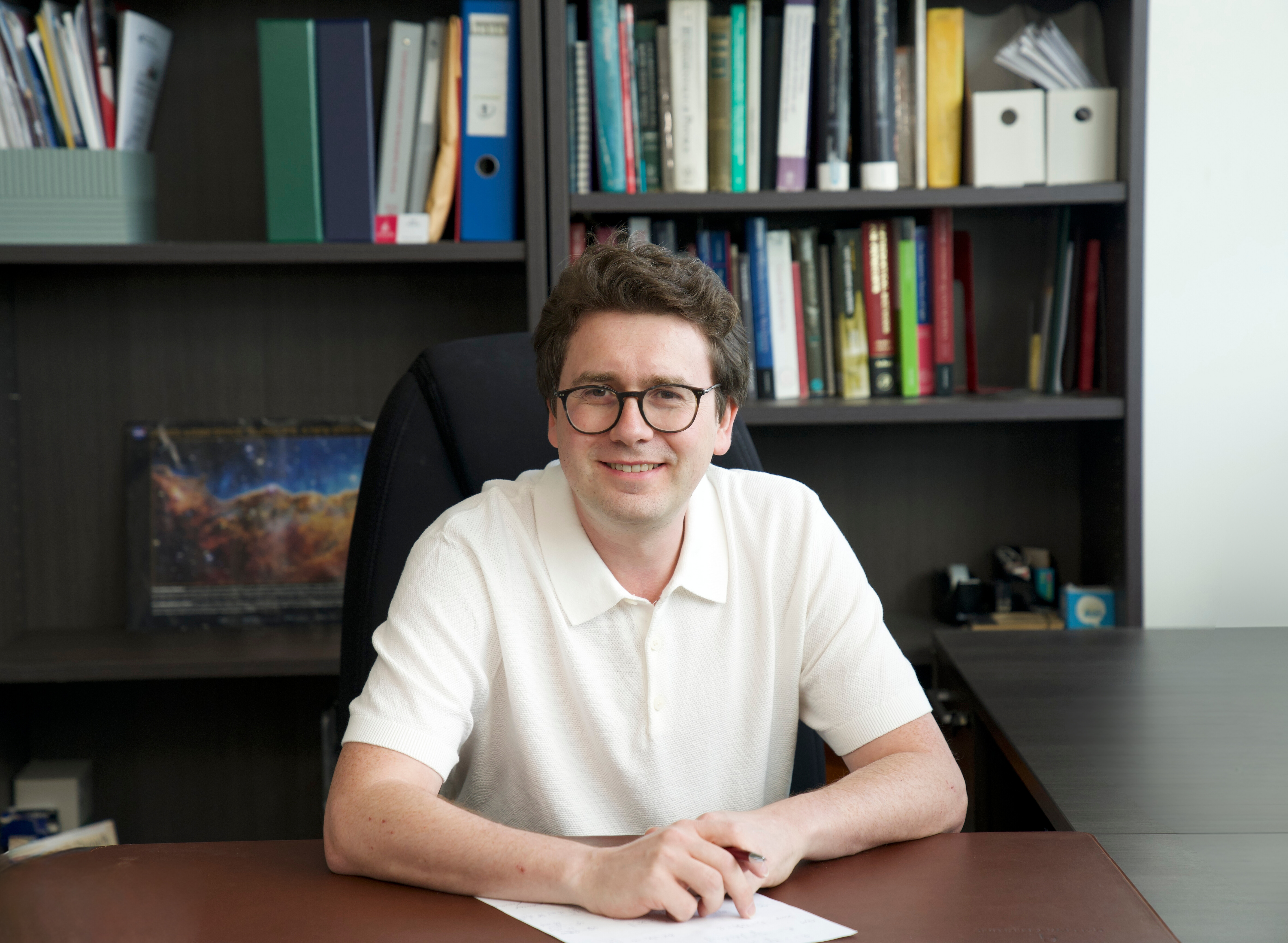 Stevens professor Igor Pikovski faces the camera directly with a pen in hand and paper on desk with a computer monitor in the background