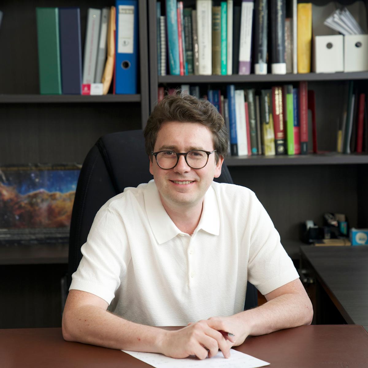 Stevens professor Igor Pikovski faces the camera directly with a pen in hand and paper on desk with a computer monitor in the background