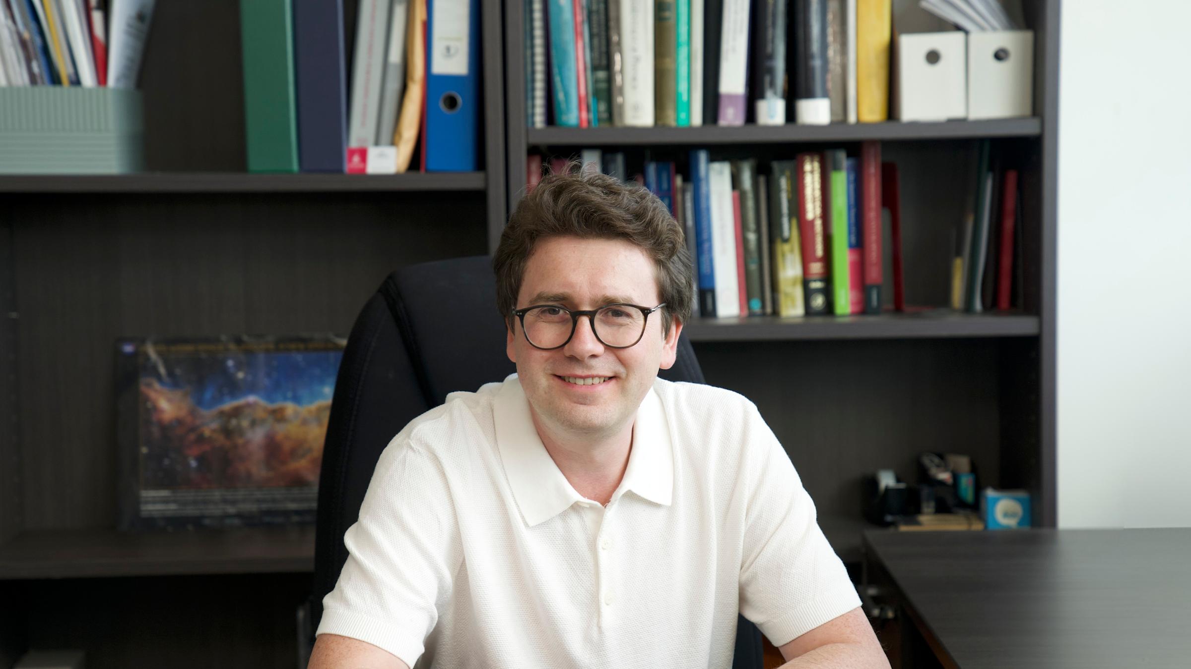 Stevens professor Igor Pikovski faces the camera directly with a pen in hand and paper on desk with a computer monitor in the background