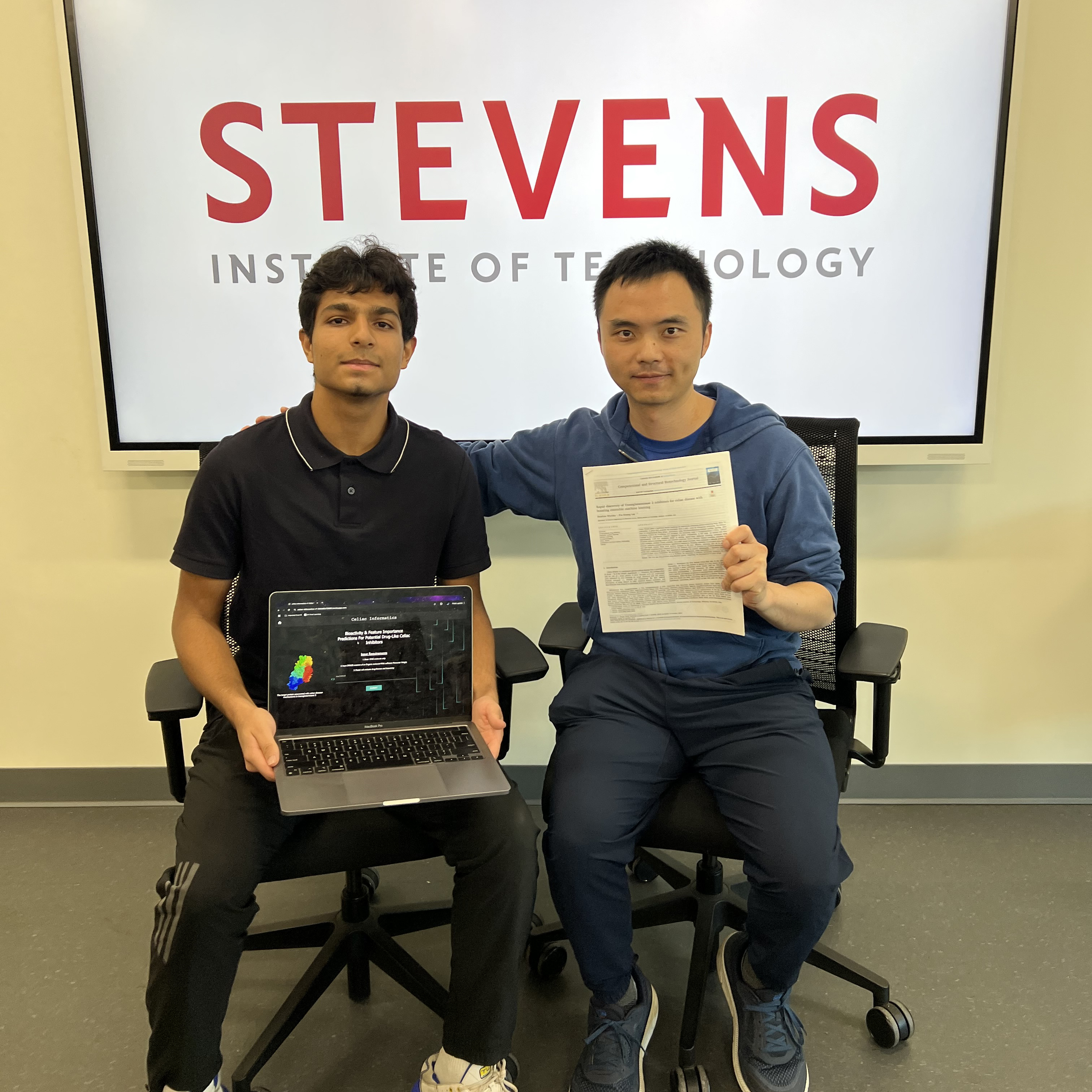 A high school student with an open laptop and Stevens professor sit in chairs in front of a white Stevens Institute of Technology sign.
