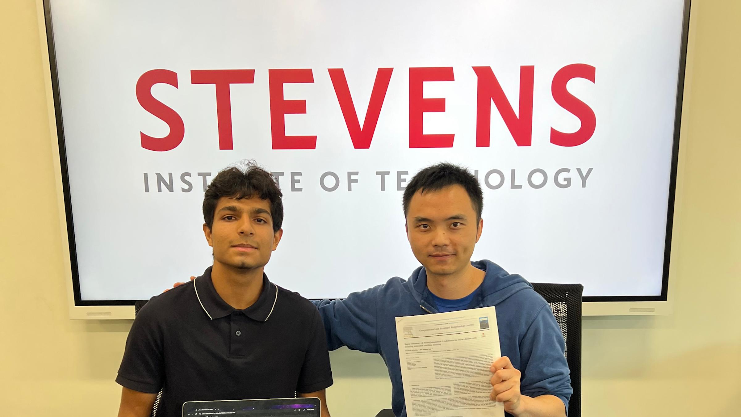 A high school student with an open laptop and Stevens professor sit in chairs in front of a white Stevens Institute of Technology sign.