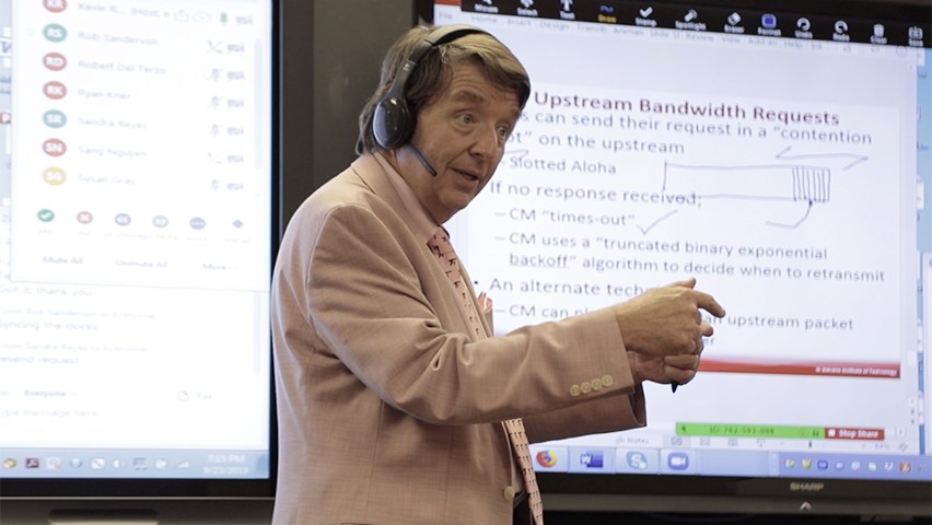 A professor talks to a physical and online class through a headset. Course materials and discussion are projected in the back.