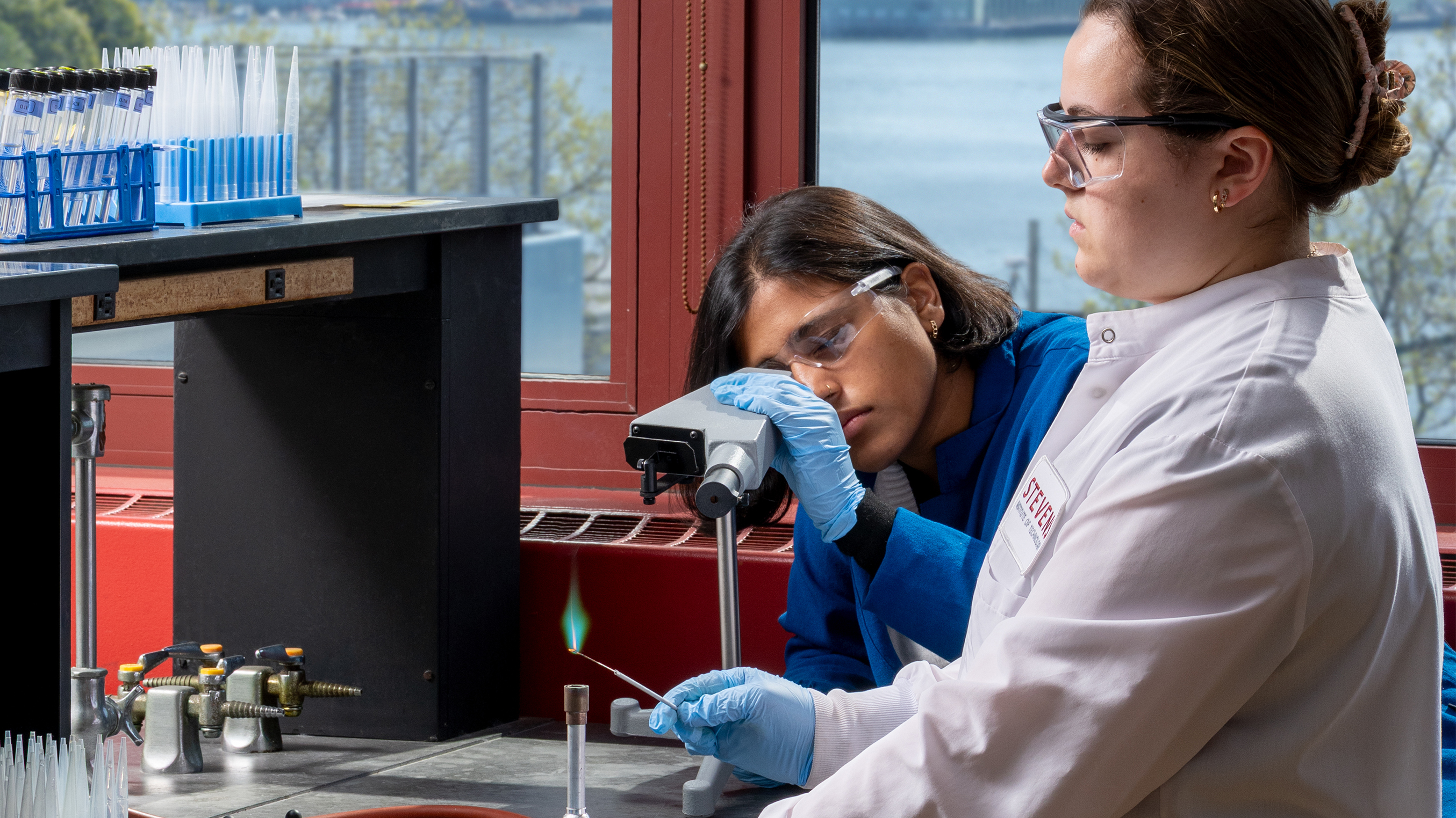 Two students in lab coats doing an experiment using a bunsen burner