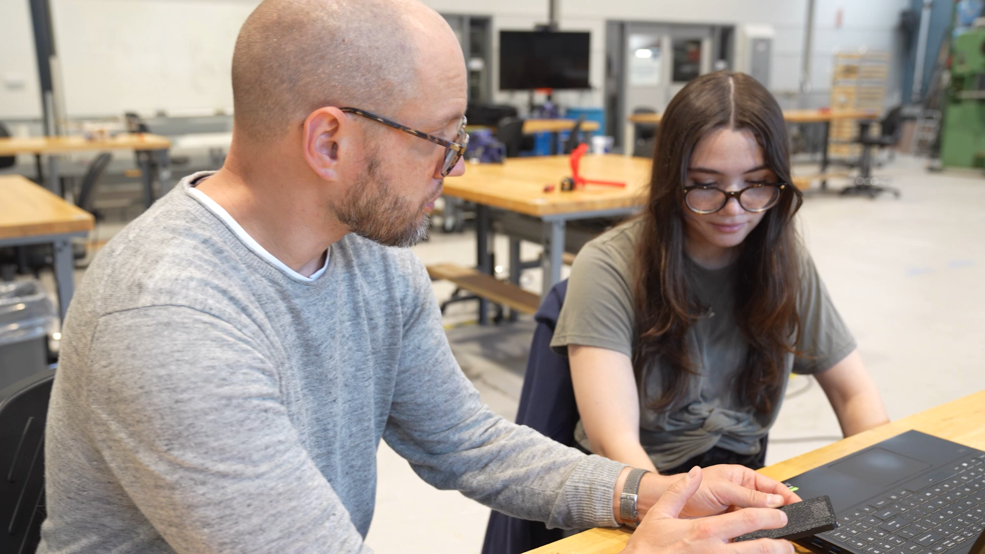 A bald man with glasses and beard in a gray sweater works with a female student wearing glasses at a laptop in a makerspace with workbenches and equipment visible in the background.