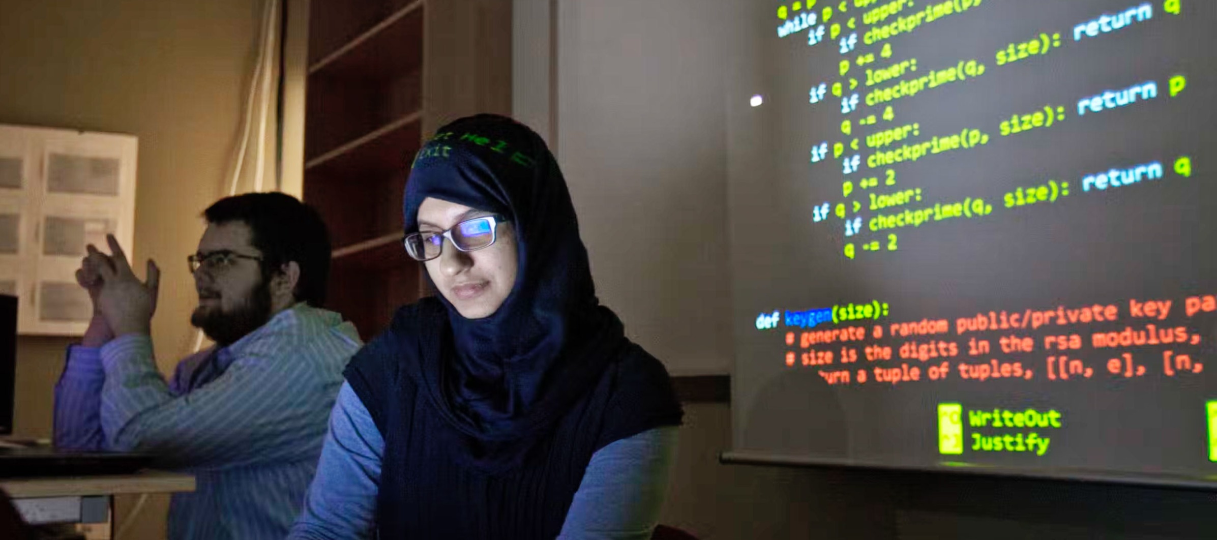 A photo of a female student wearing a hijab working in a dimly lit computer lab with code on the screen in the background