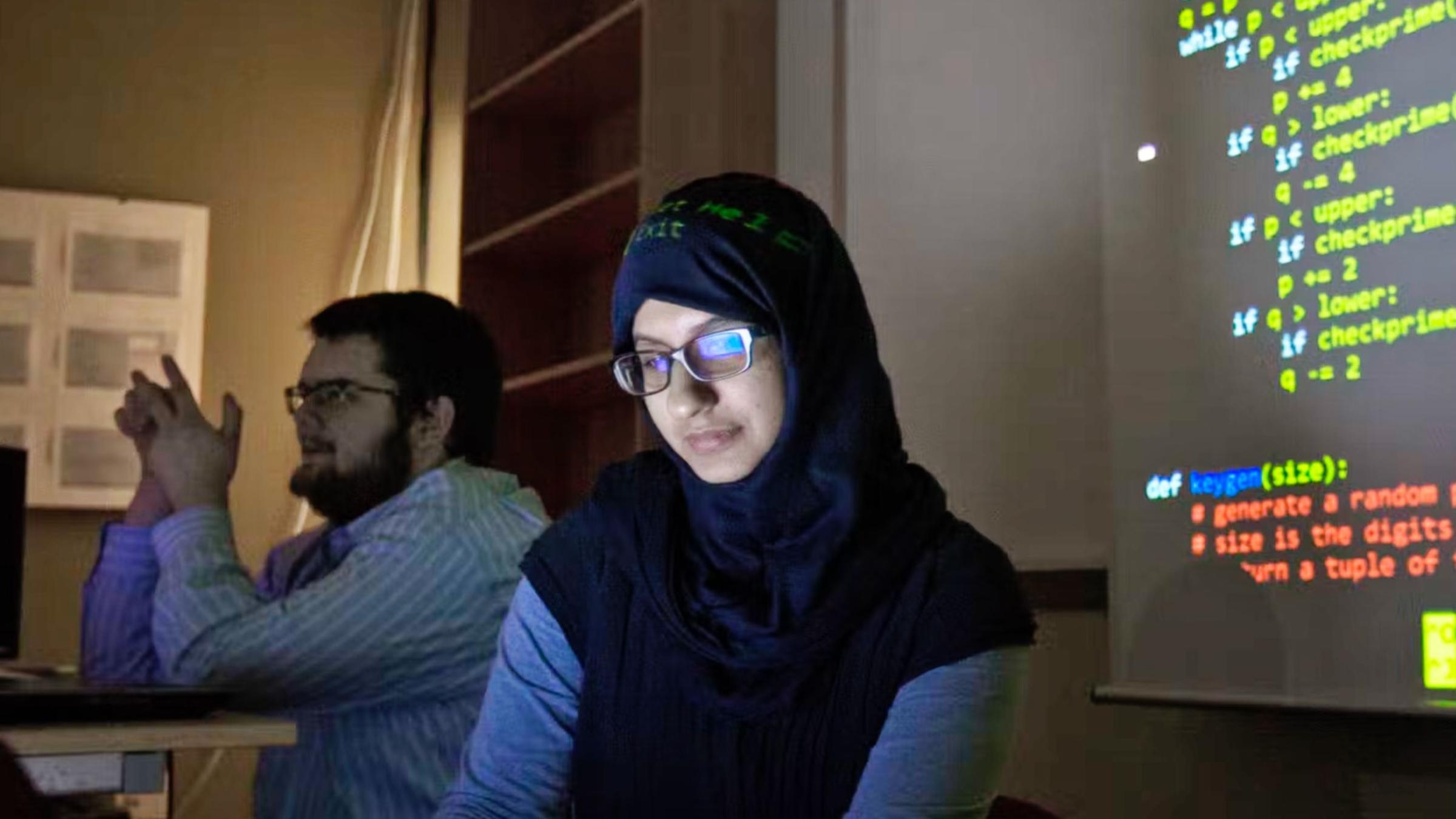A photo of a female student wearing a hijab working in a dimly lit computer lab with code on the screen in the background