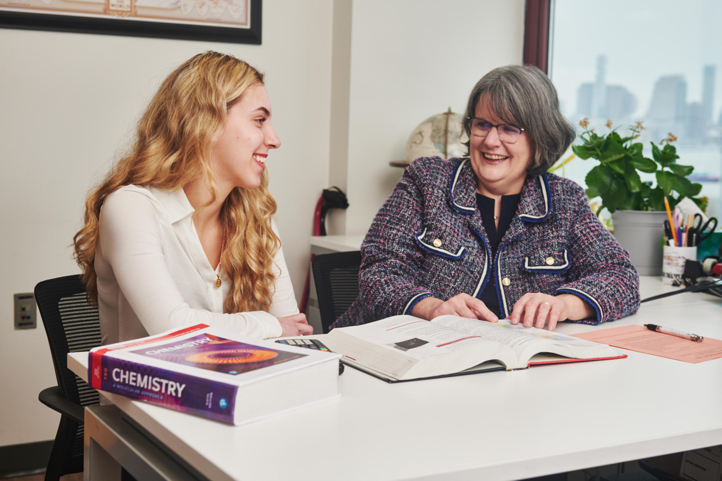Two women in a Stevens campus office. Two chemistry textbooks are on the desk, with a window partly visible in the background.