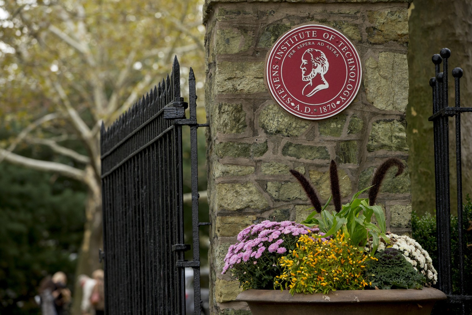an image of the gatehouse, with Stevens red emblem. Pink, white and green flowers are at the bottom.