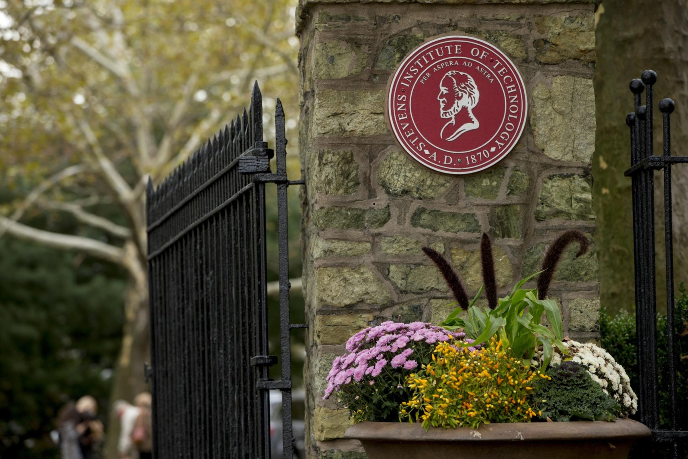an image of the gatehouse, with Stevens red emblem. Pink, white and green flowers are at the bottom.