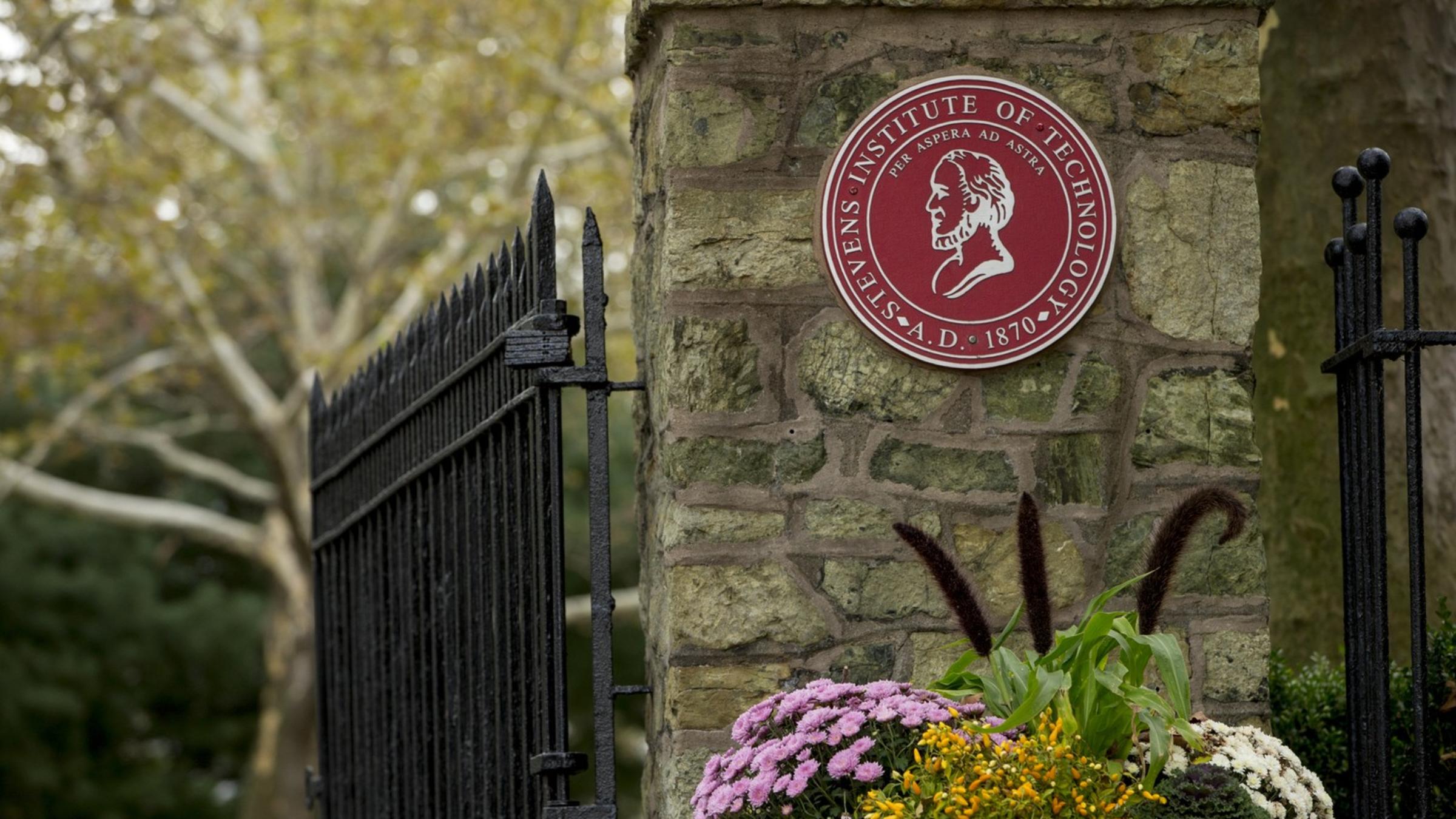 an image of the gatehouse, with Stevens red emblem. Pink, white and green flowers are at the bottom.