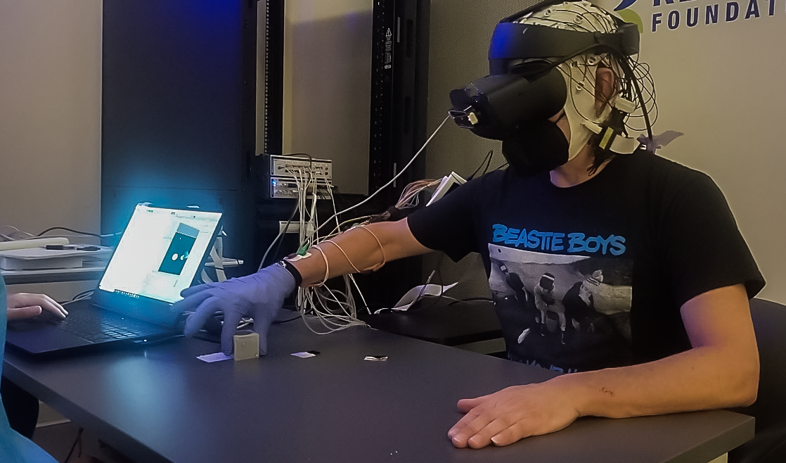 A male in a black shirt and black VR headset picks up an object on a black table with a gloved hand.