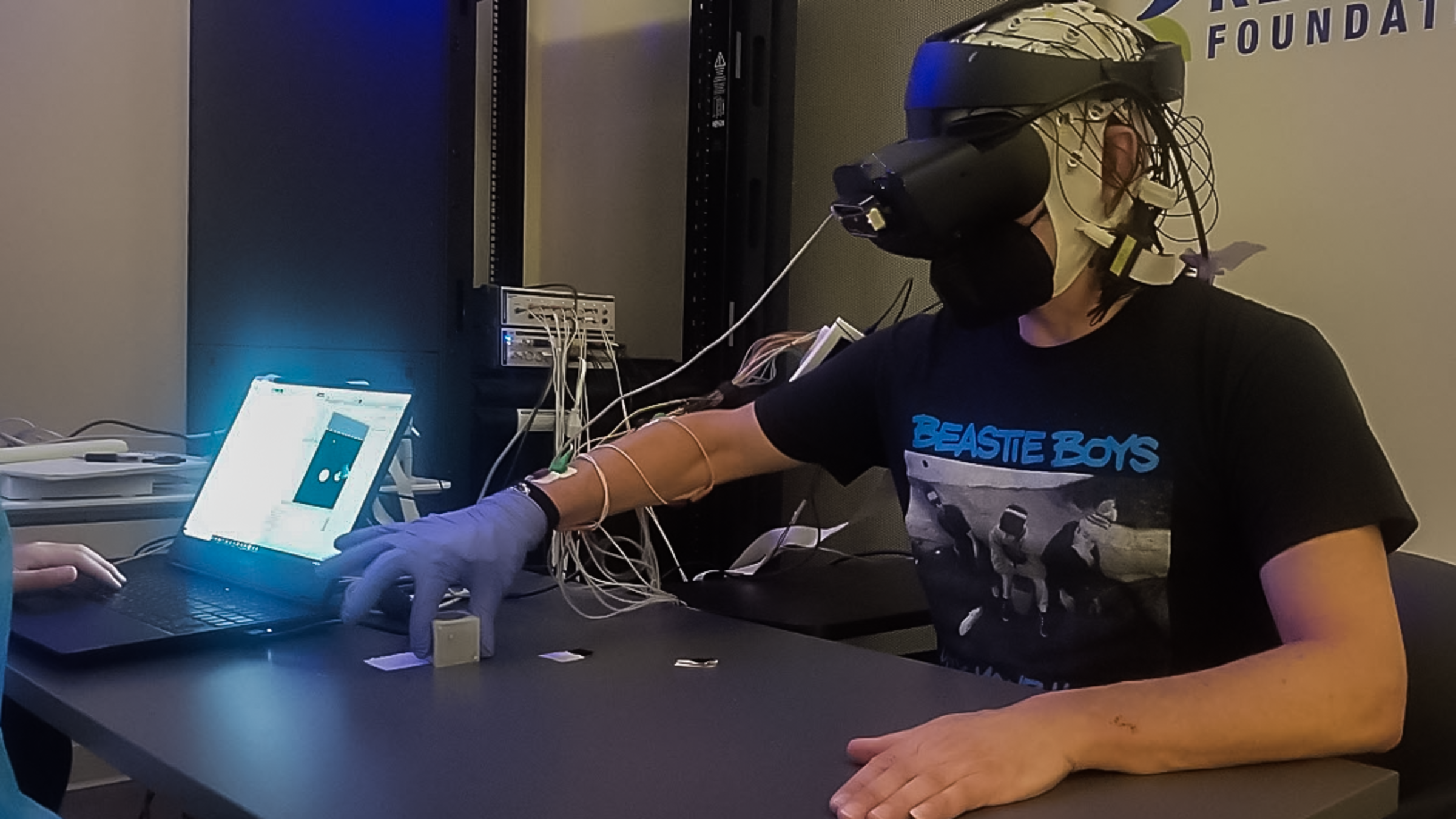 A male in a black shirt and black VR headset picks up an object on a black table with a gloved hand.