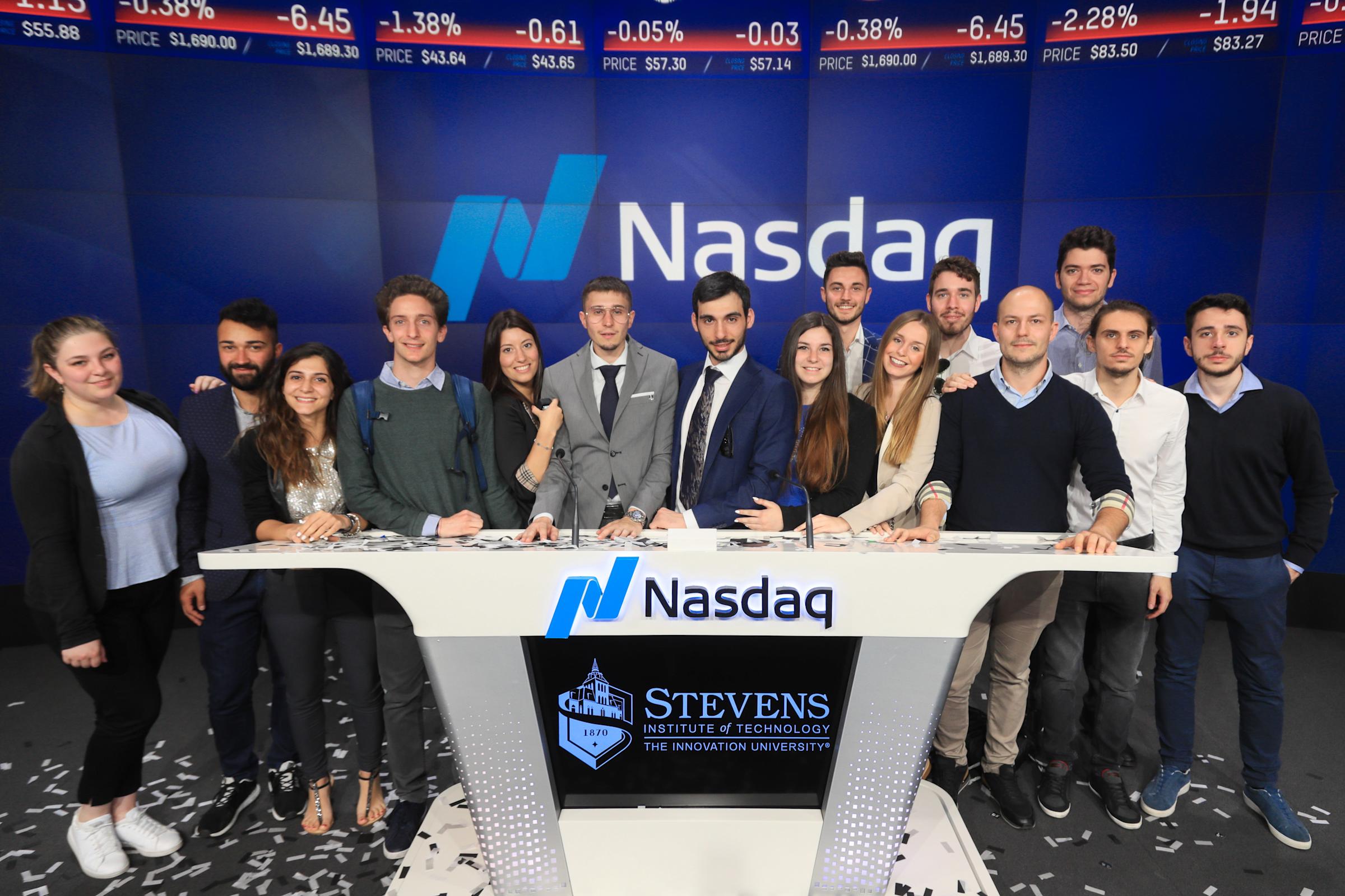 Stevens Institute of Technology students and faculty gather at the Nasdaq trading desk studio, standing behind the white Nasdaq podium with the Stevens logo displayed. Stock market tickers scroll on screens above the blue Nasdaq-branded backdrop.
