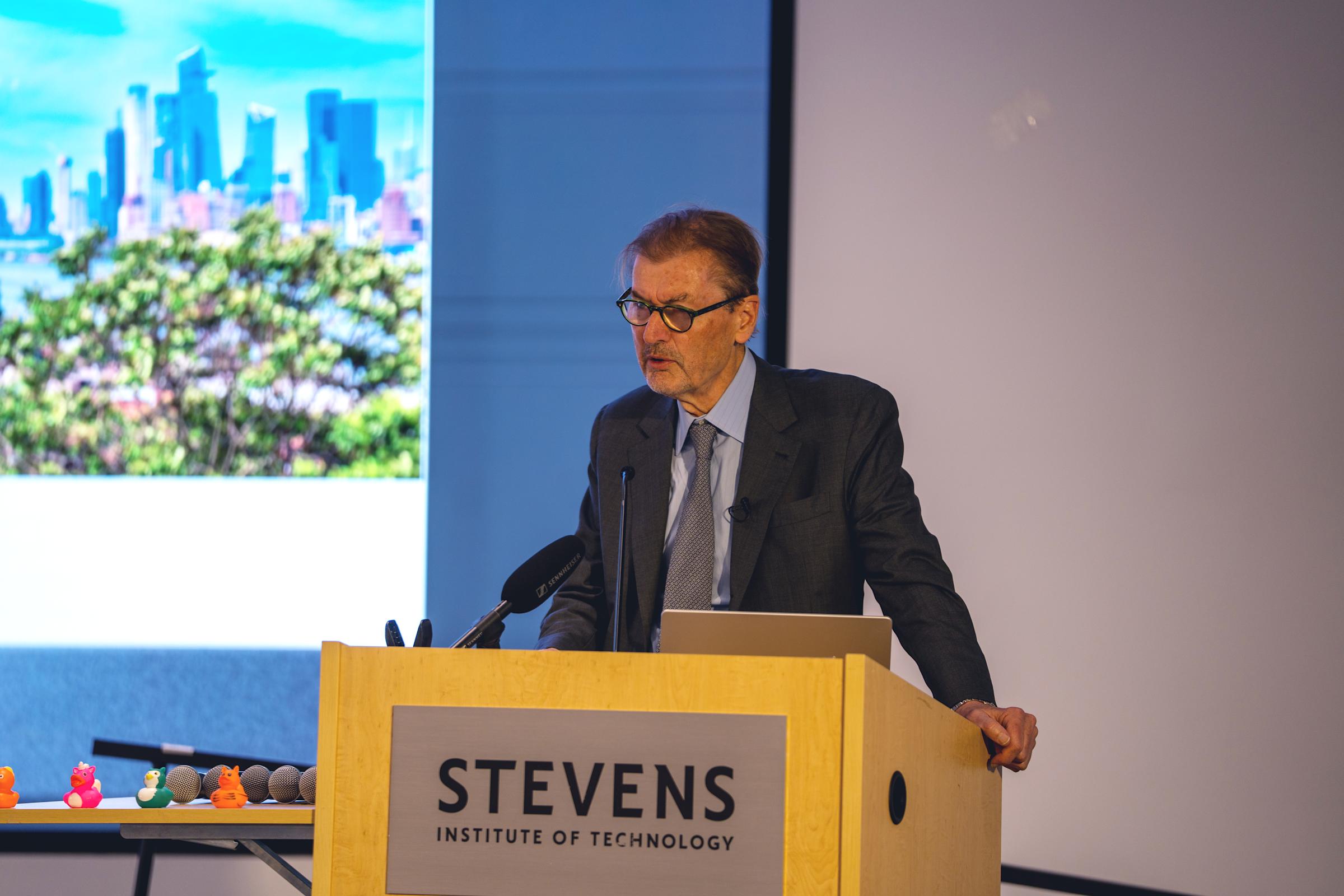 Man in dark suit and tie with glasses stands at wooden Stevens Institute of Technology podium with microphone, presenting with city skyline visible on projection screen behind him. Colorful rubber ducks visible at edge of stage.
