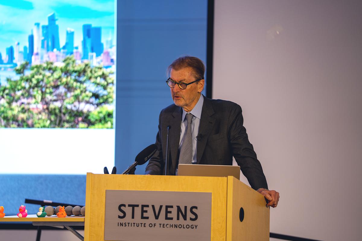 Man in dark suit and tie with glasses stands at wooden Stevens Institute of Technology podium with microphone, presenting with city skyline visible on projection screen behind him. Colorful rubber ducks visible at edge of stage.