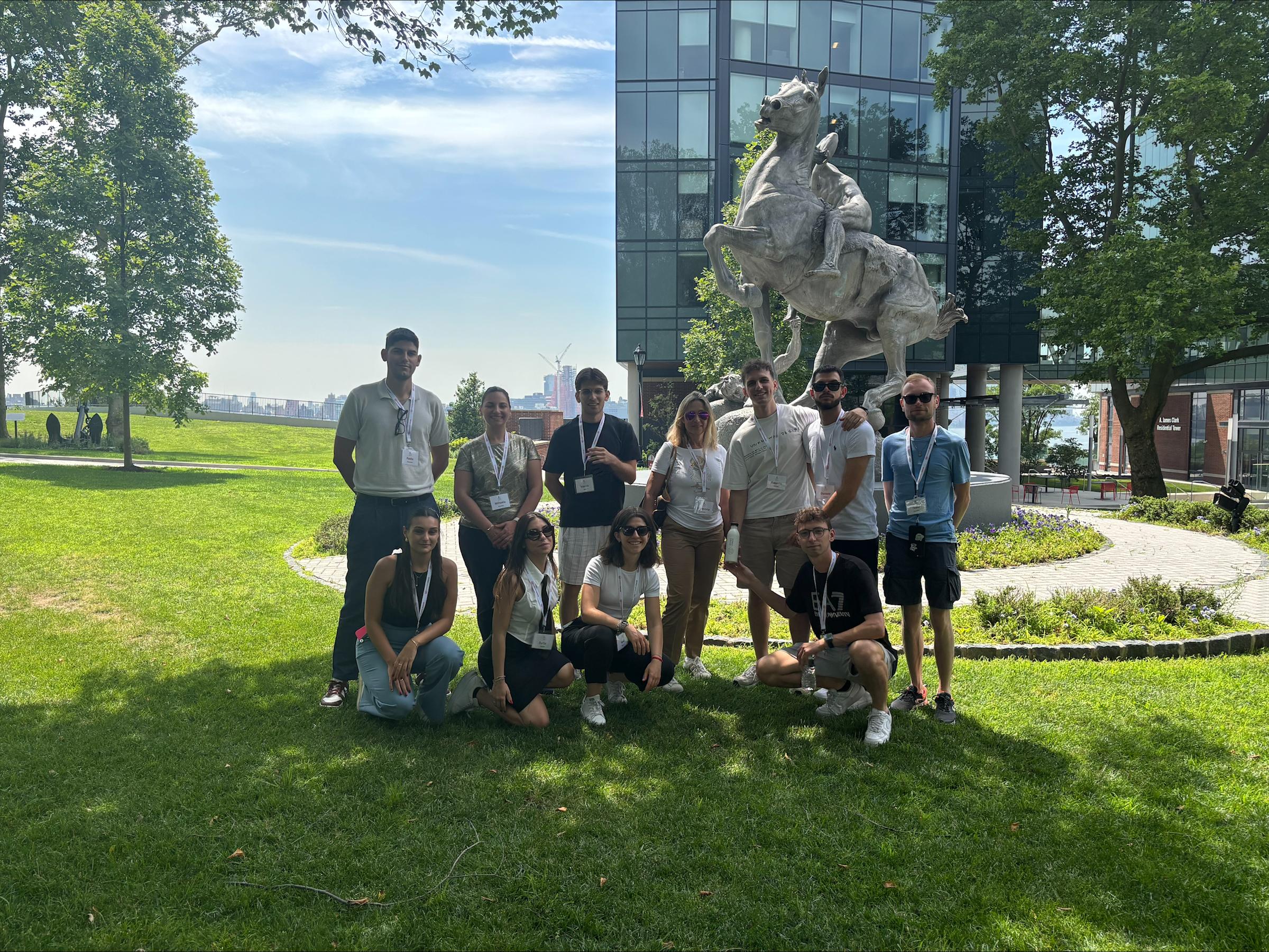 A group of young professionals wearing name badges pose on a grassy lawn in front of a large bronze statue and modern glass building, with the Manhattan skyline and water visible in the background.