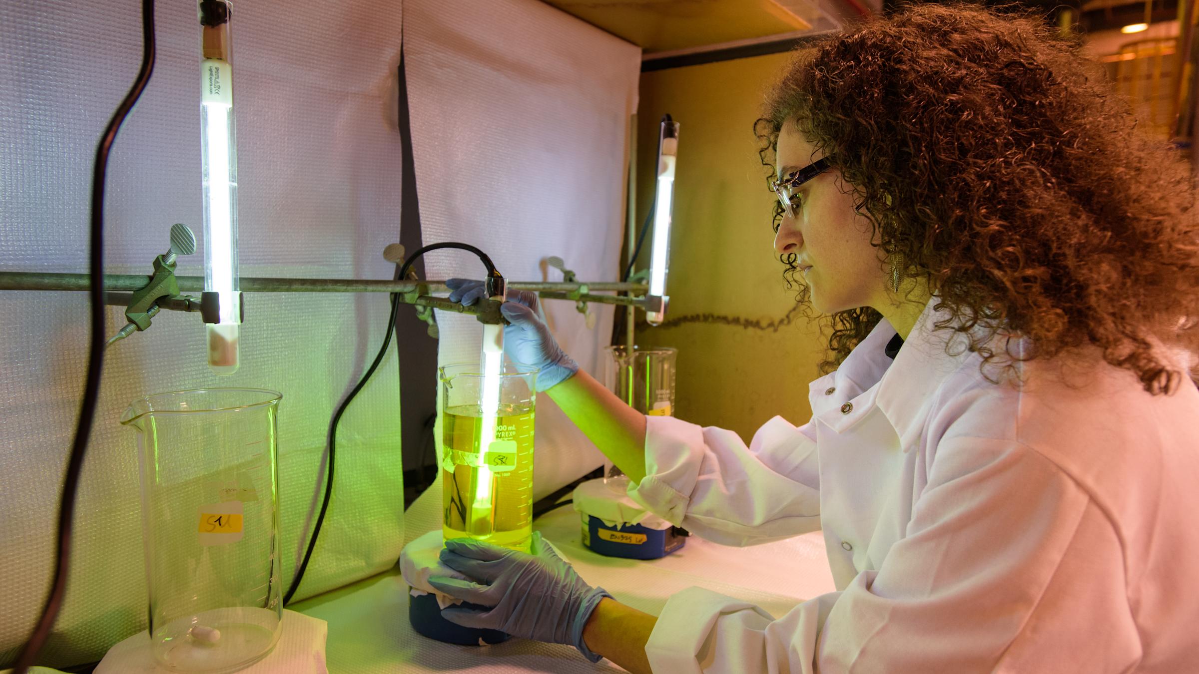 A student in a lab coat and gloves works with light and fluids in glass beakers within a environmental engineering lab.