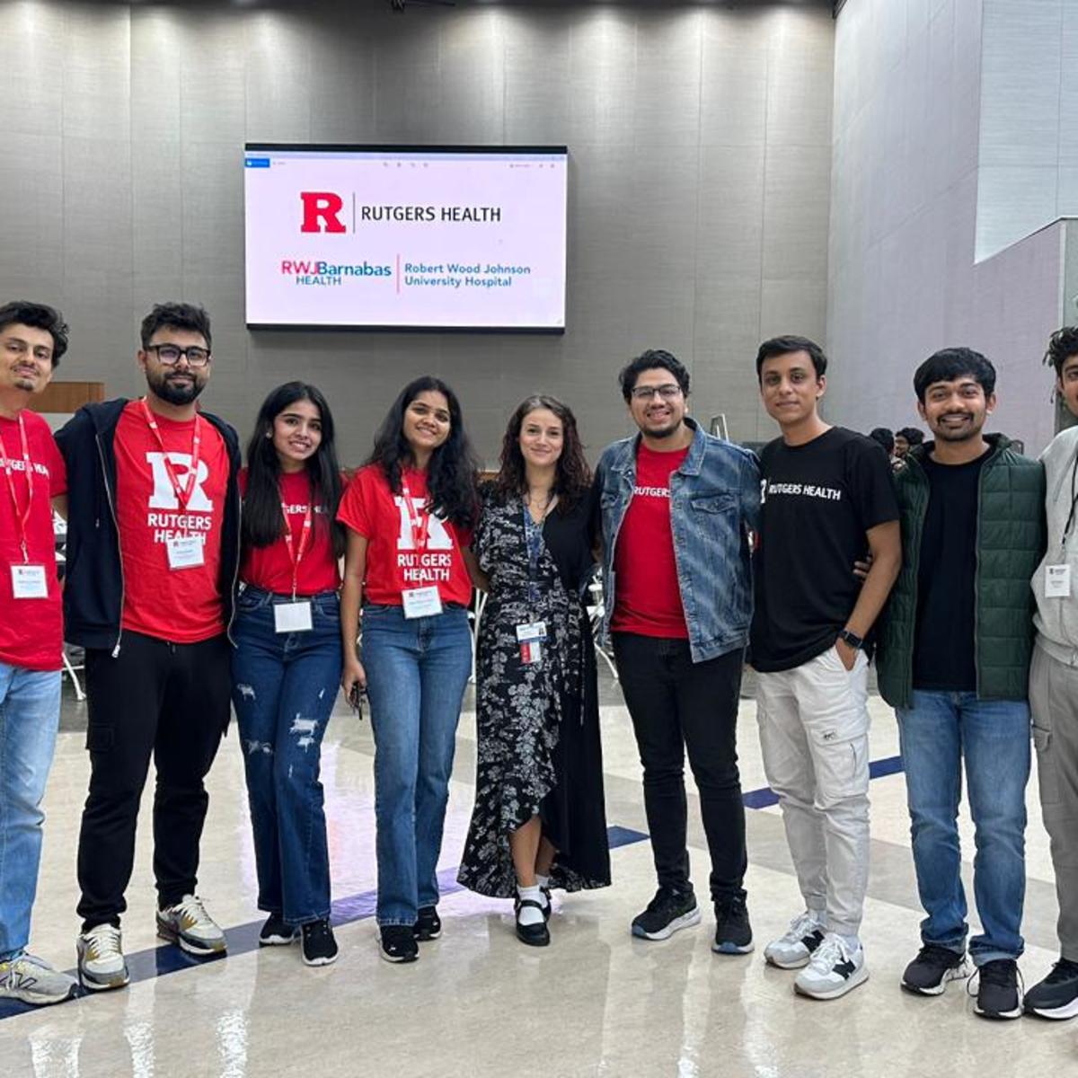 Nine students and young professionals pose in a modern hospital lobby in front of a Rutgers Health display. Several wear red Rutgers Health t-shirts and conference badges. The group stands in The Arlene and Henry Schwartzman Courtyard.