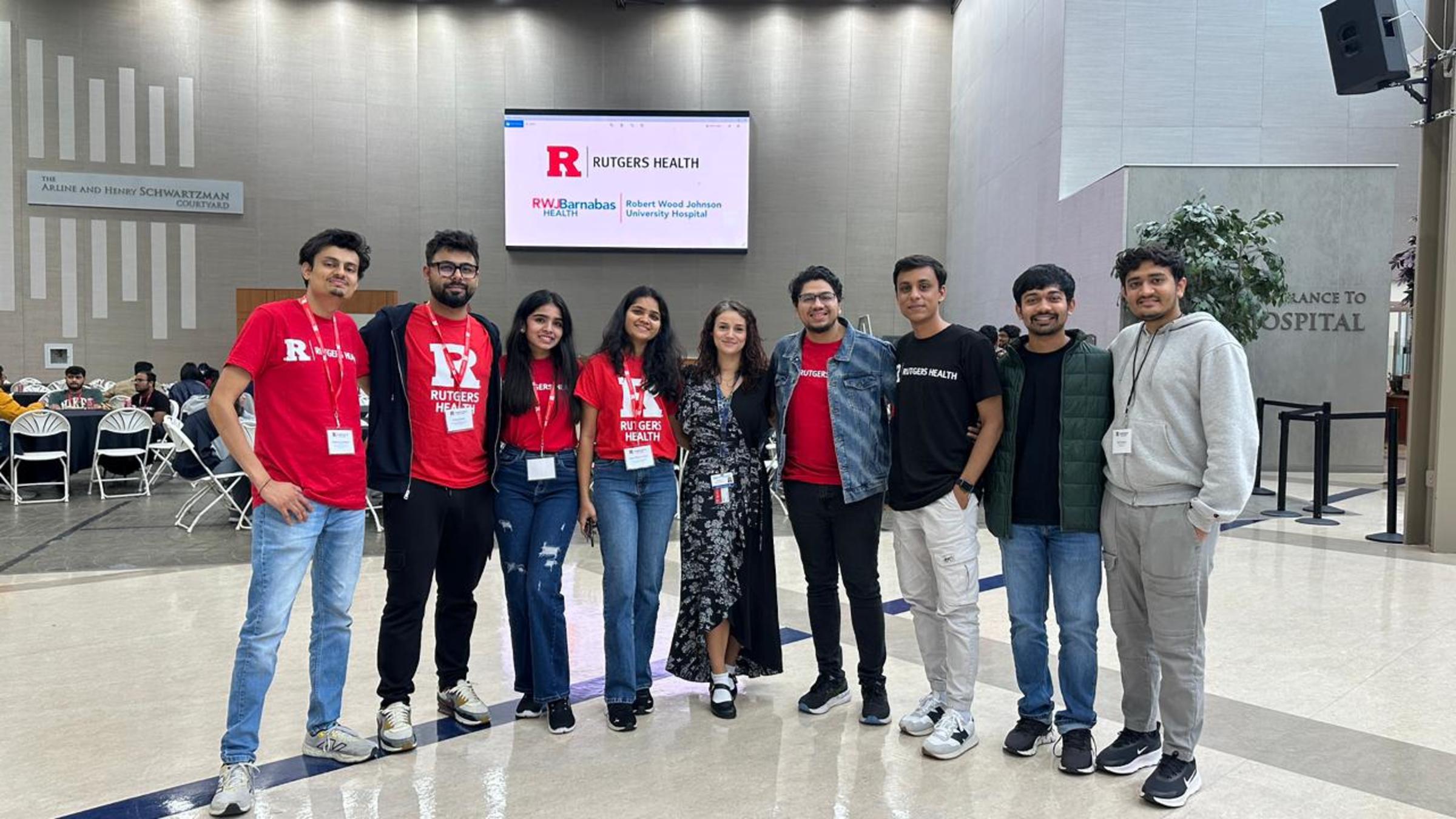 Nine students and young professionals pose in a modern hospital lobby in front of a Rutgers Health display. Several wear red Rutgers Health t-shirts and conference badges. The group stands in The Arlene and Henry Schwartzman Courtyard.