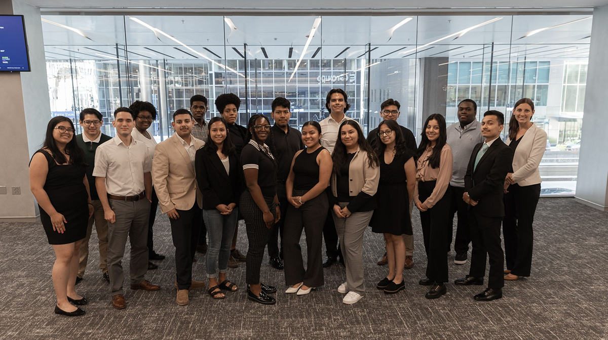 Group of a dozen college students take a photo together in room with floor to ceiling glass windows