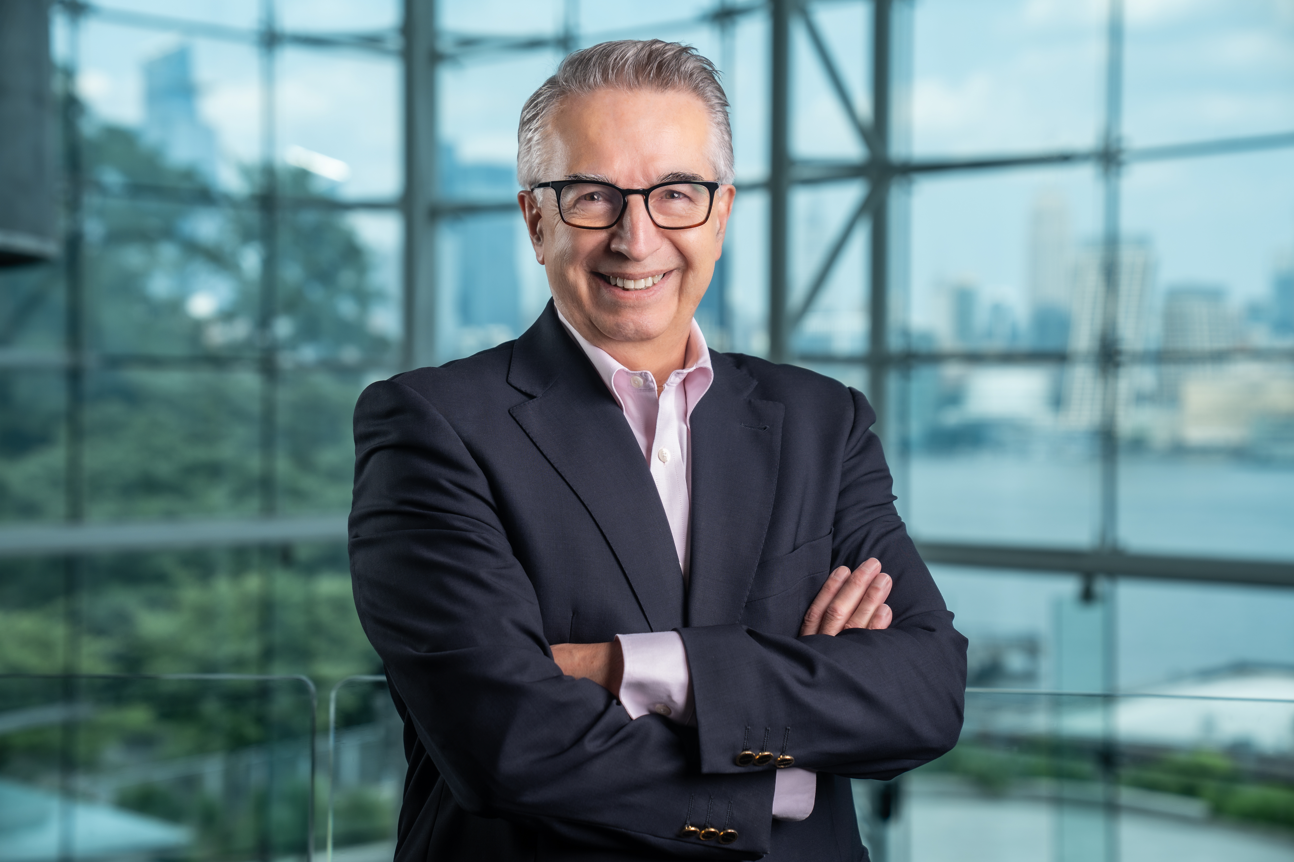 Gregory Prastacos in a navy blue blazer with his arms folded and the New York City skyline in the background through the Babbio Center windows.