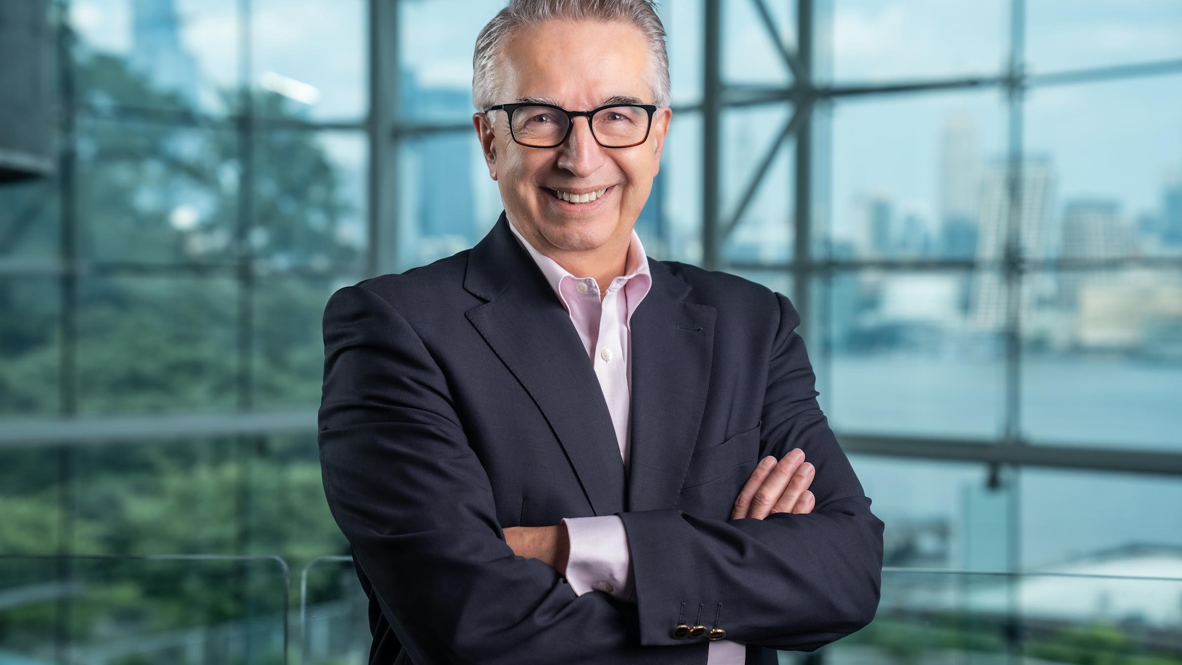 Gregory Prastacos in a navy blue blazer with his arms folded and the New York City skyline in the background through the Babbio Center windows.