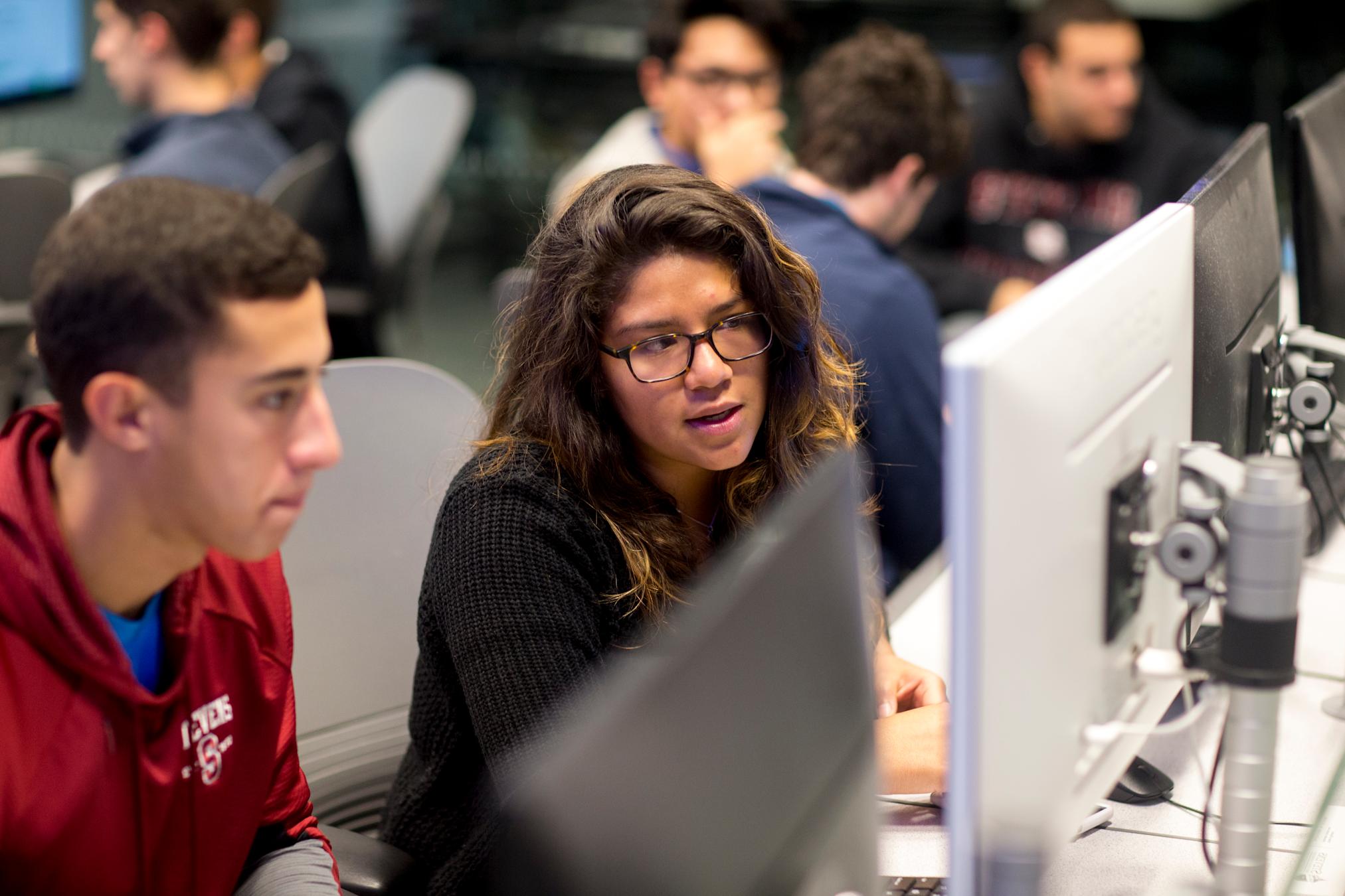 Student gestures to monitor while speaking as another student looks on curiously