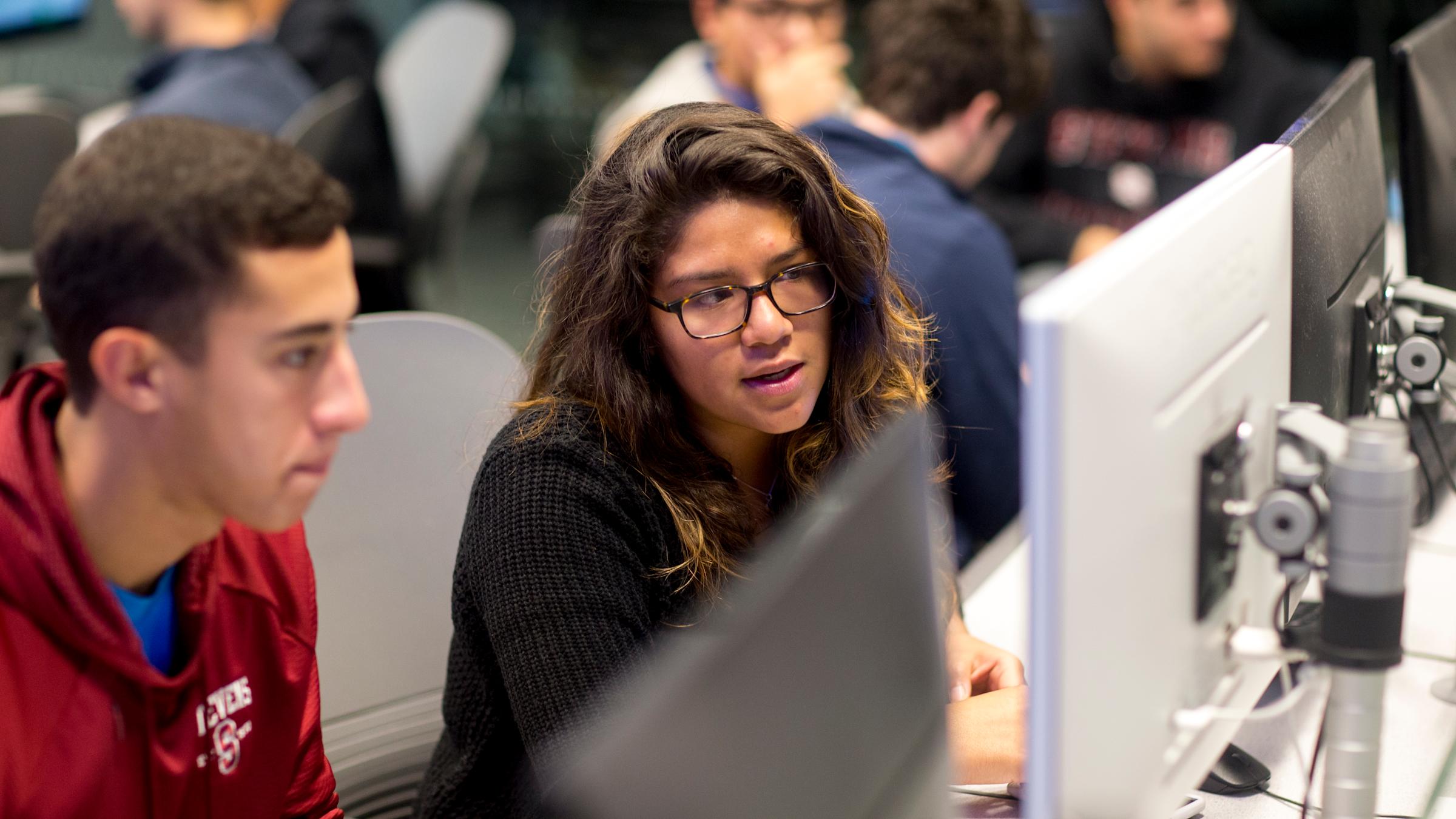 Student gestures to monitor while speaking as another student looks on curiously