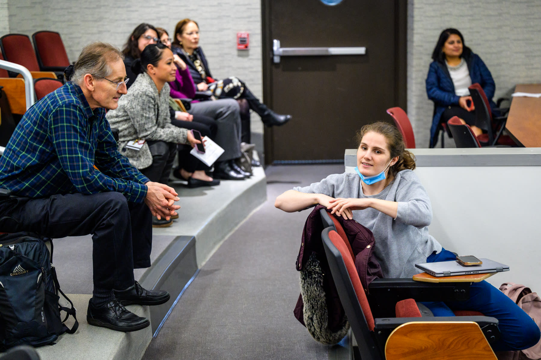 Group of people sitting in chairs in an auditorium, with a woman and man in the foreground speaking to each other