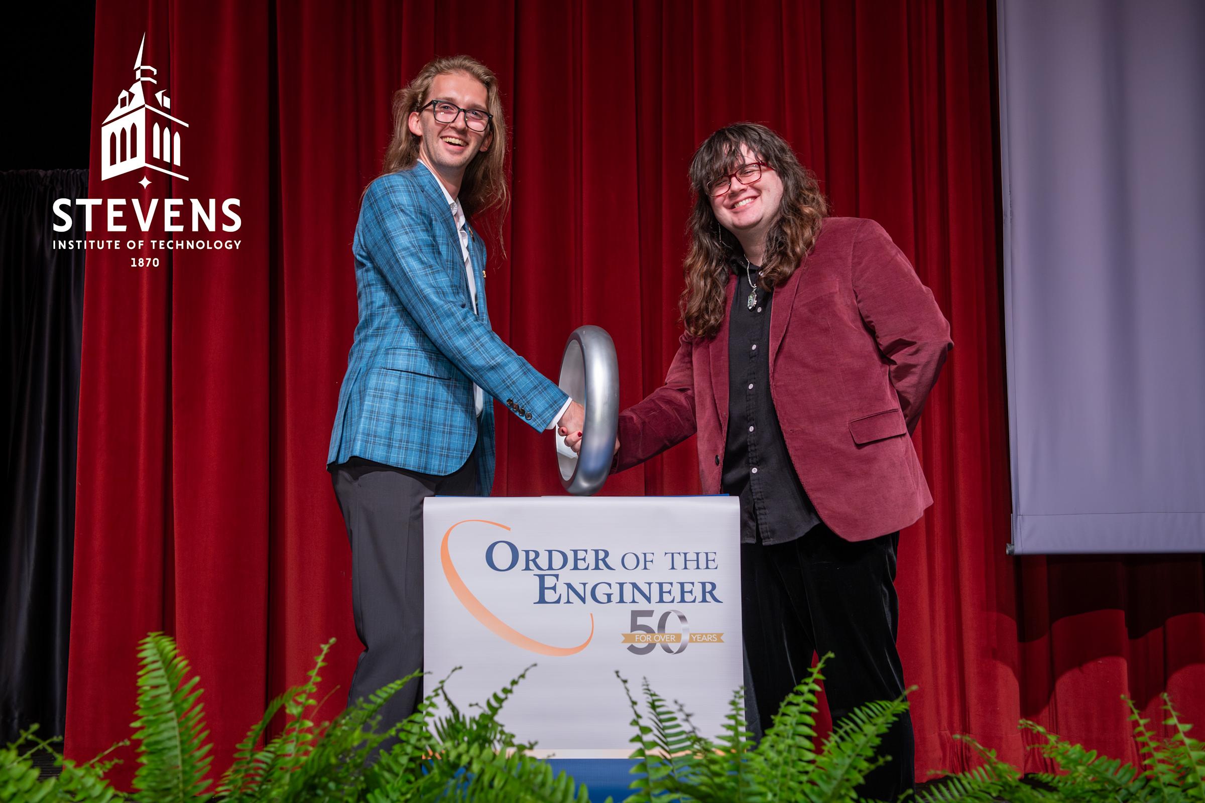 Two people stand behind a podium wearing business clothes. On the podium it reads "Order of the Engineer." The two people are shaking hands through a large ring that is on the podium.