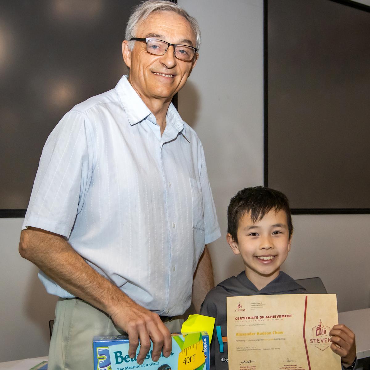 Stevens professor Pavel Dubovski, an adult male, left, poses with a young male participant holding an award from the 2025 Stevens Math Olympiad.