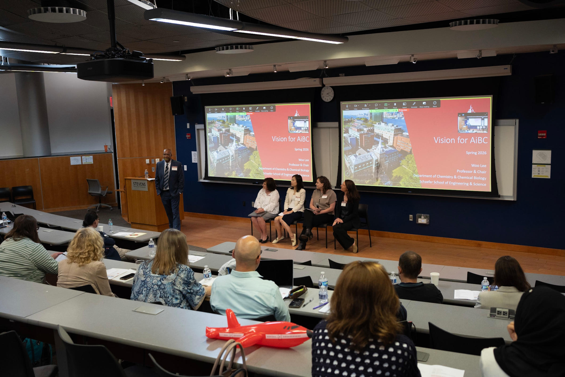 This photo points at the front of the lecture hall where there are four women sitting in chairs in front of the projectors with a man standing to the left who is introducing them and the powerpoint in the background reads Vision for AiBC.