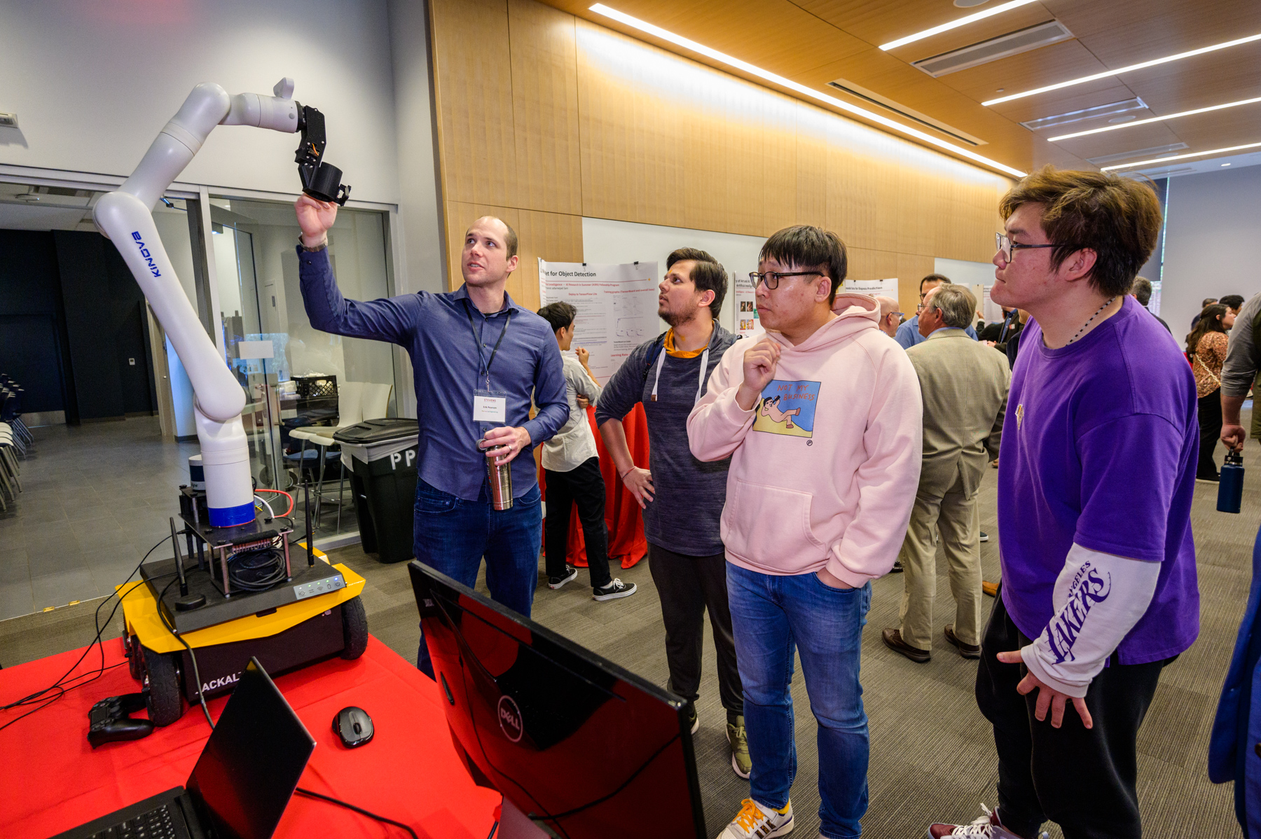 A professor demonstrates a robotic arm to a group of students at the 2023 SIAI Relaunch Event, with posters and laptops set up on tables nearby.