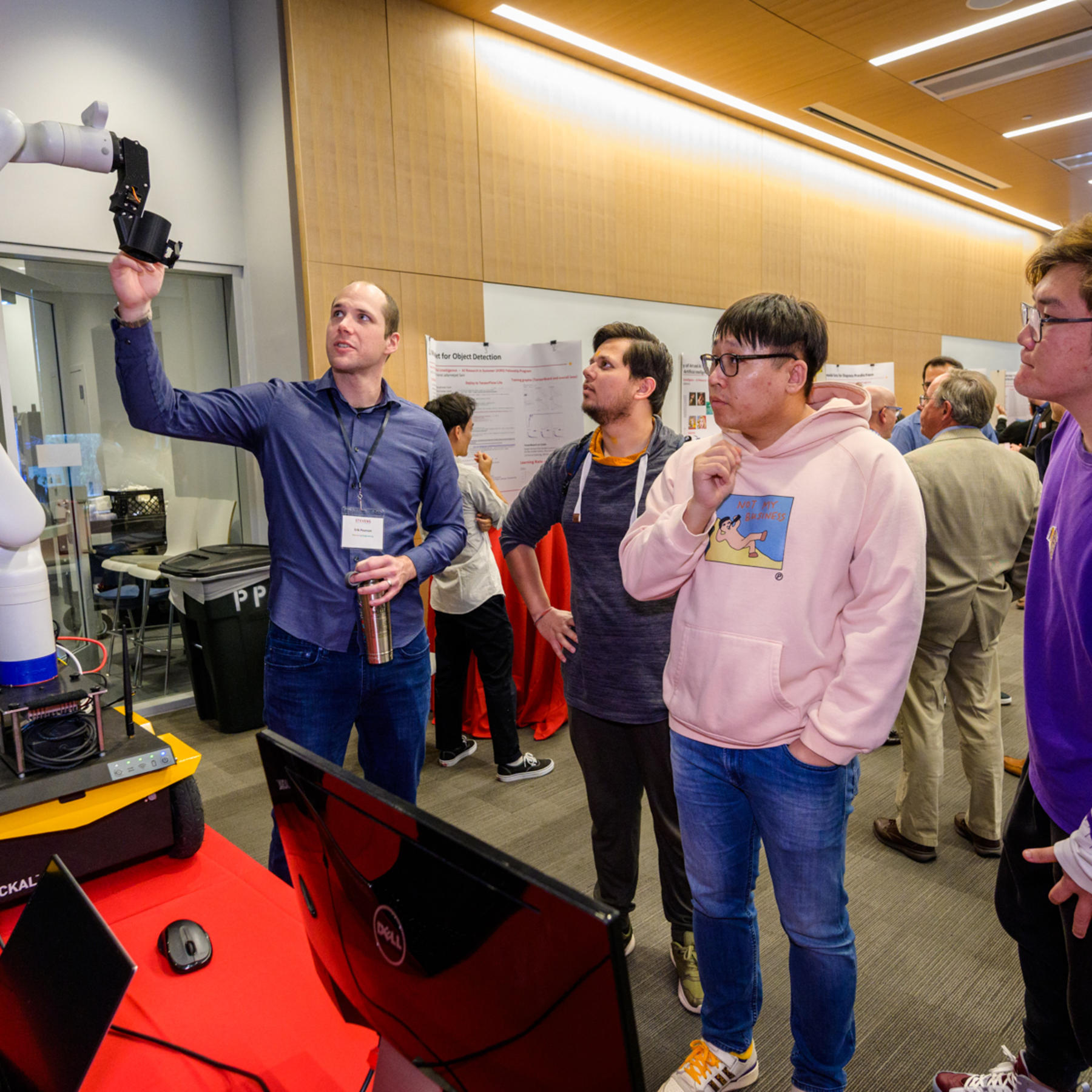A professor demonstrates a robotic arm to a group of students at the 2023 SIAI Relaunch Event, with posters and laptops set up on tables nearby.