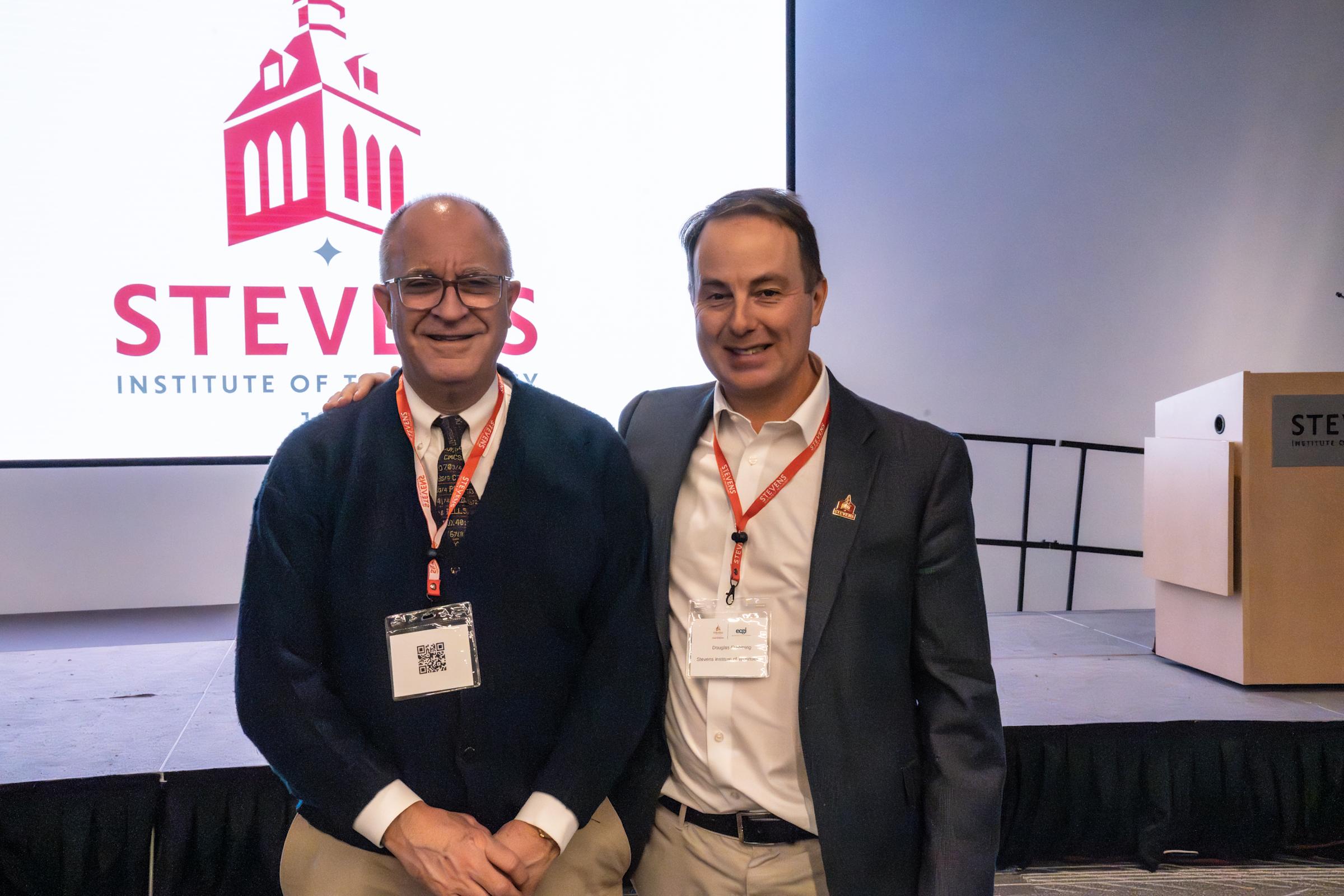 Two men in business attire wearing red lanyards and conference badges pose together smiling in front of a large Stevens Institute of Technology logo backdrop at what appears to be an academic conference or event.