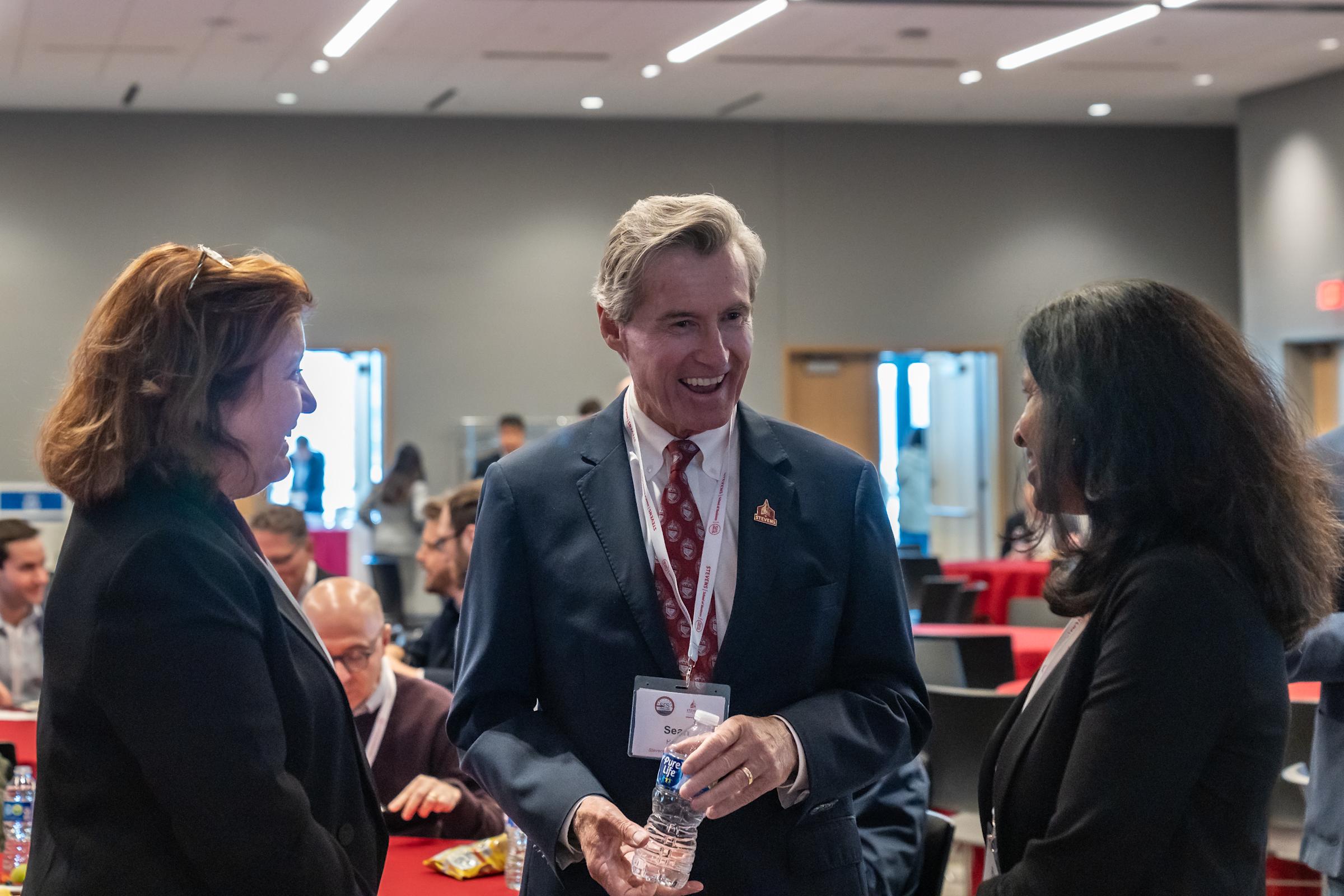 Three professionals network at a conference, with a smiling man in a dark suit and red tie holding a water bottle at the center, flanked by two women in dark attire, all wearing conference badges in a modern venue.