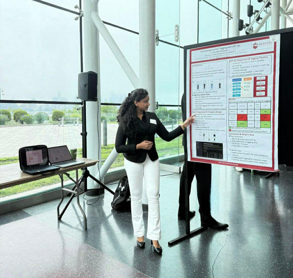 A woman with curly dark hair in a black blazer and white pants presents her research poster at an academic conference. She stands next to a large Stevens Institute poster with charts and data, in a modern glass-walled venue with laptops on a nearby table.