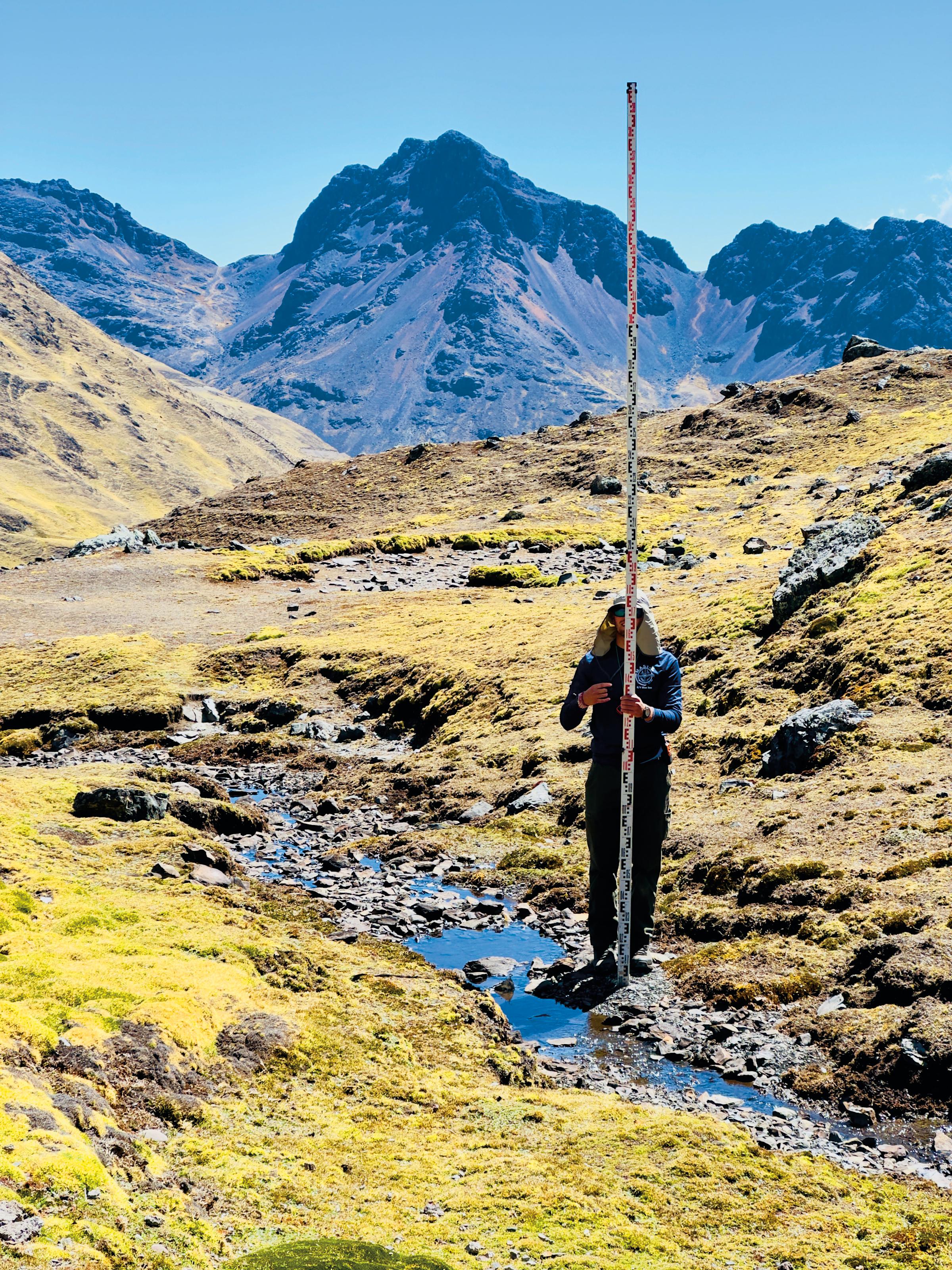 A student holding a tall ruler takes measurements on a hillside.