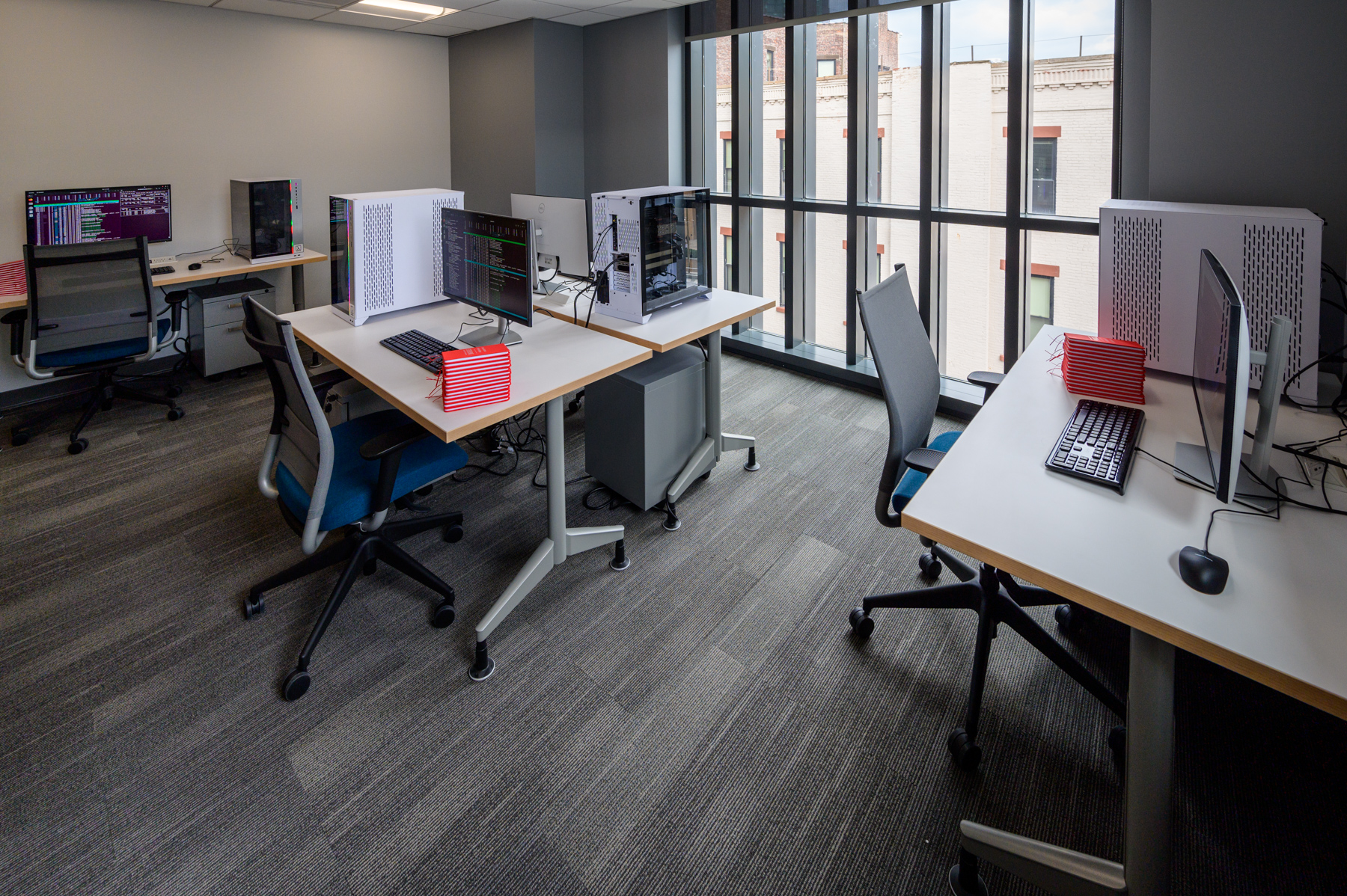 Modern workspace with multiple workstations featuring dual monitors, desktop computers, ergonomic office chairs and red notebooks. 