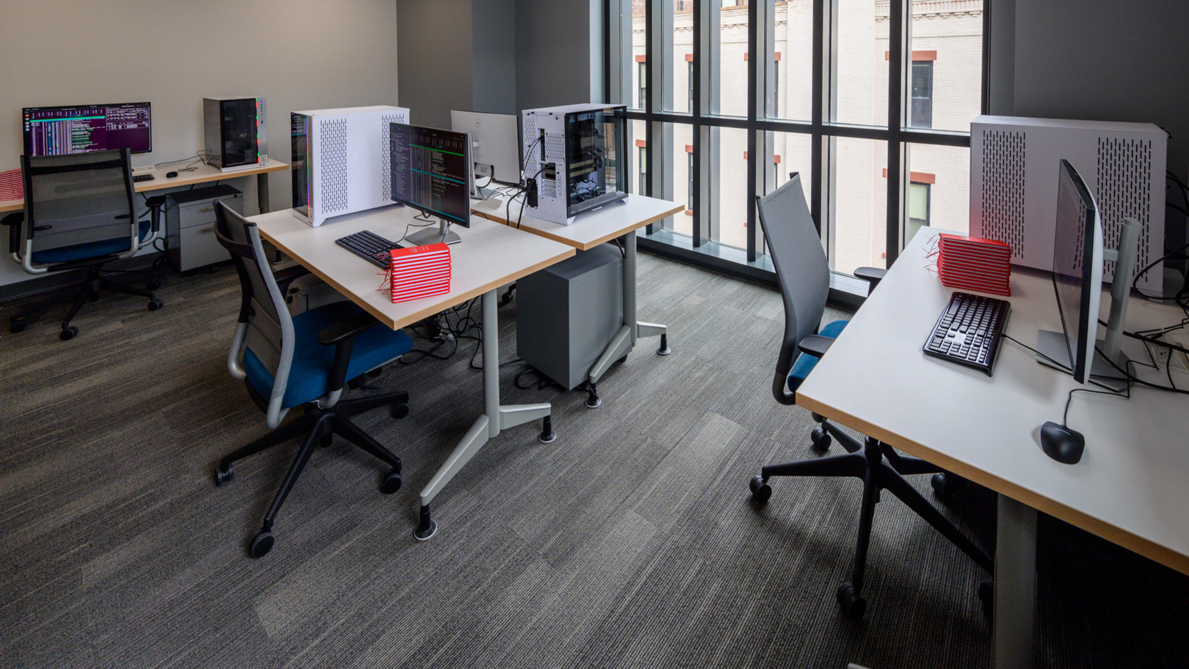 Modern workspace with multiple workstations featuring dual monitors, desktop computers, ergonomic office chairs and red notebooks.