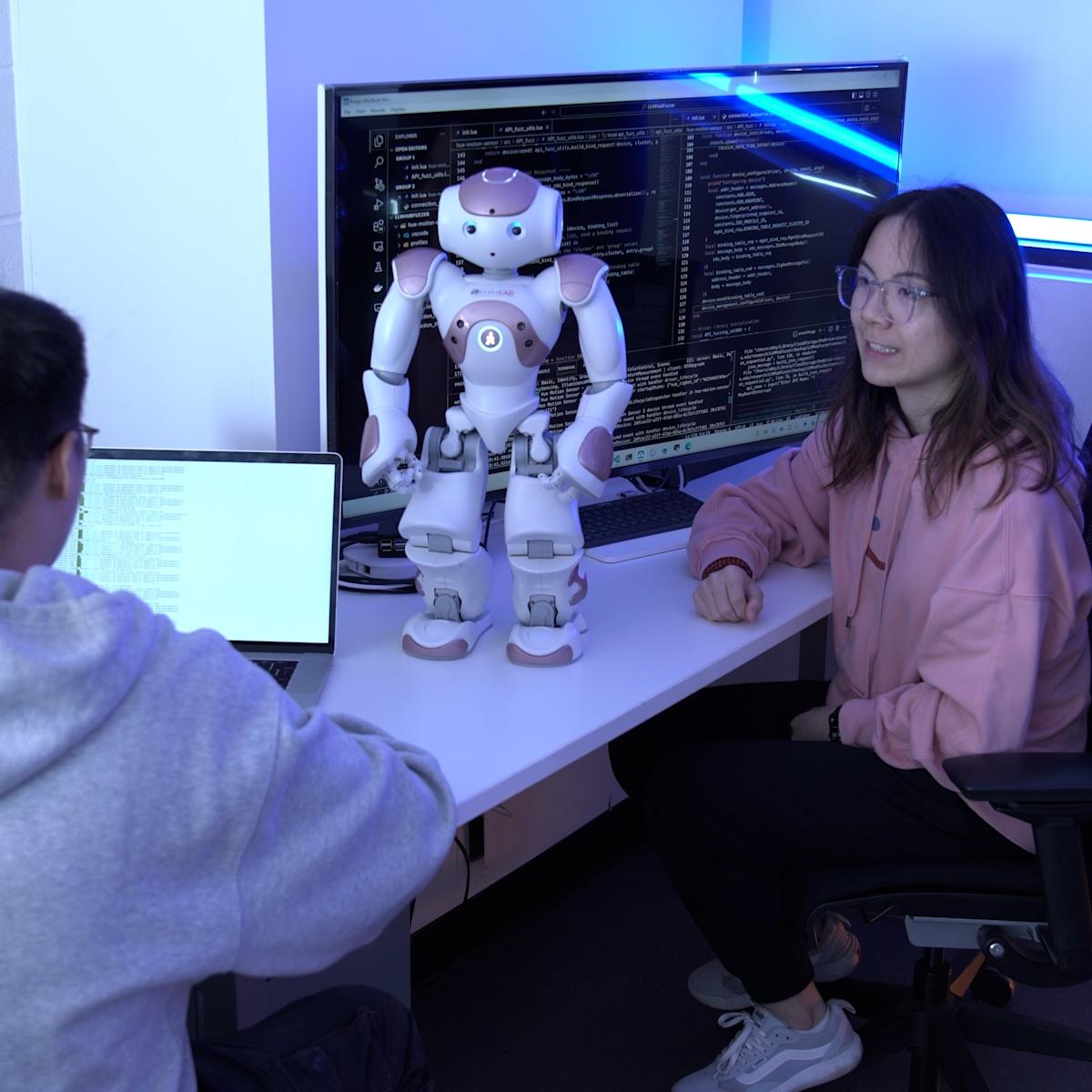 Two students sit at a desk with a standing robot.